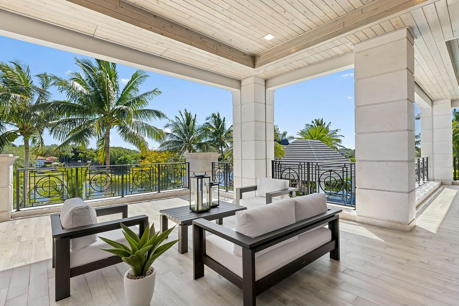 This image showcases a luxurious outdoor balcony or patio area, furnished with modern seating arrangements including a sofa and chairs with white cushions and dark frames. The space is framed by elegant columns and a decorative railing, offering a view of lush palm trees and a serene body of water. The flooring appears to be light-colored wood-look tile, contributing to the sophisticated and relaxing ambiance.