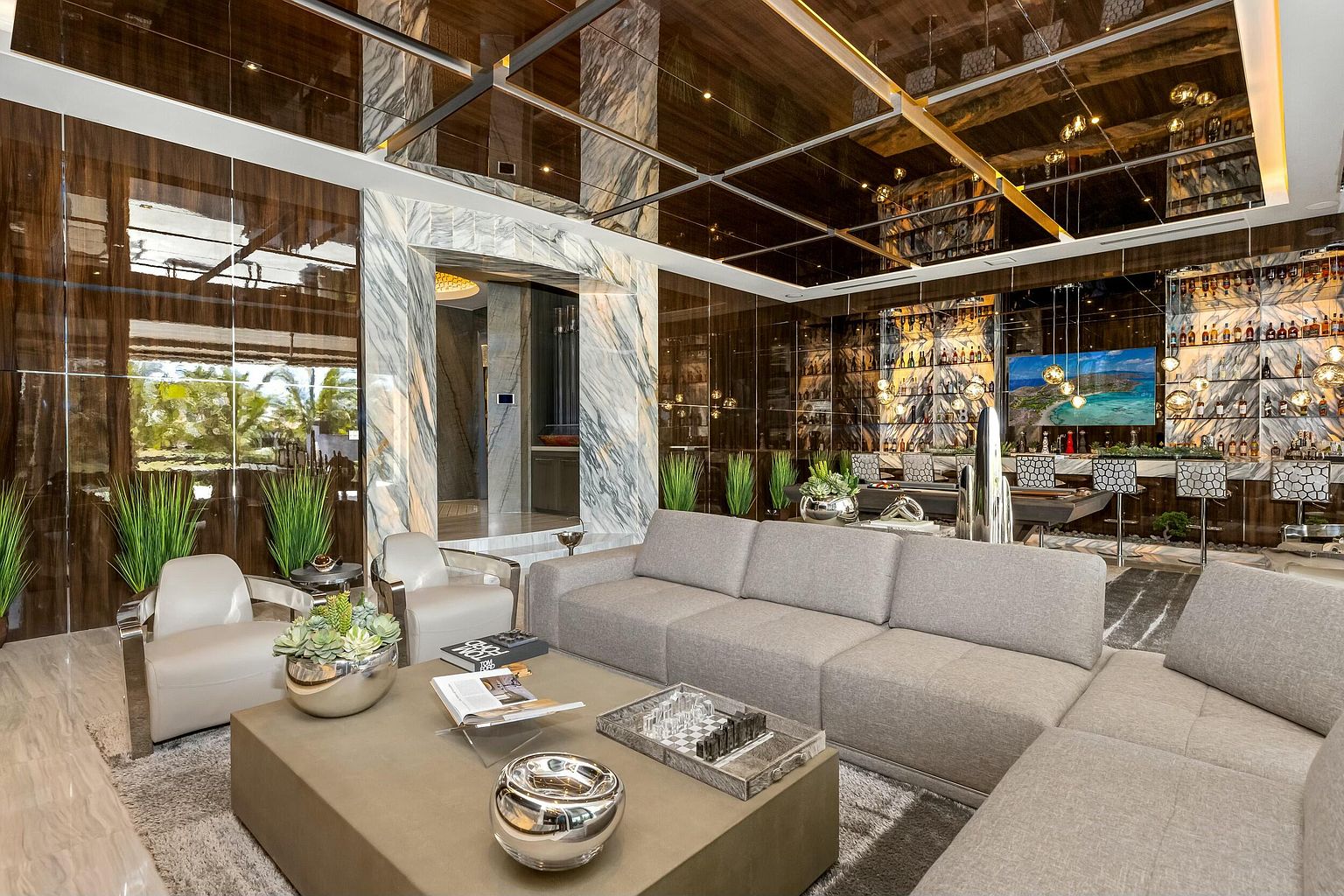 This is a luxurious living room featuring a large sectional sofa, a modern coffee table with decorative objects, and stylish armchairs. The room boasts high-end finishes, including mirrored ceilings, marble accents, and a fully stocked bar area in the background. The overall impression is one of opulence and sophisticated design.
