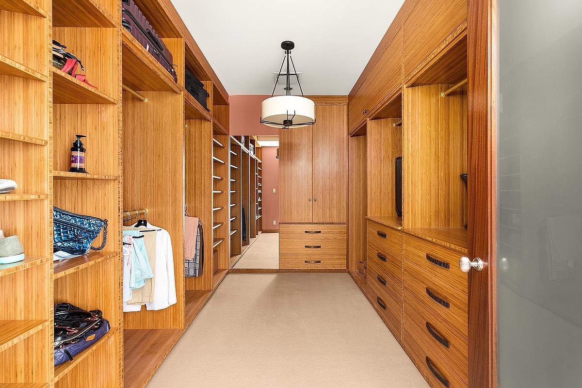 This spacious walk-in closet features warm, custom-built wood cabinetry that provides extensive shelving, hanging space, and integrated drawers. The symmetrical layout, centered by a classic chandelier, creates an organized and luxurious atmosphere. A neutral carpeted floor and a mirrored back wall enhance the sense of depth and elegance in this well-appointed storage space.
