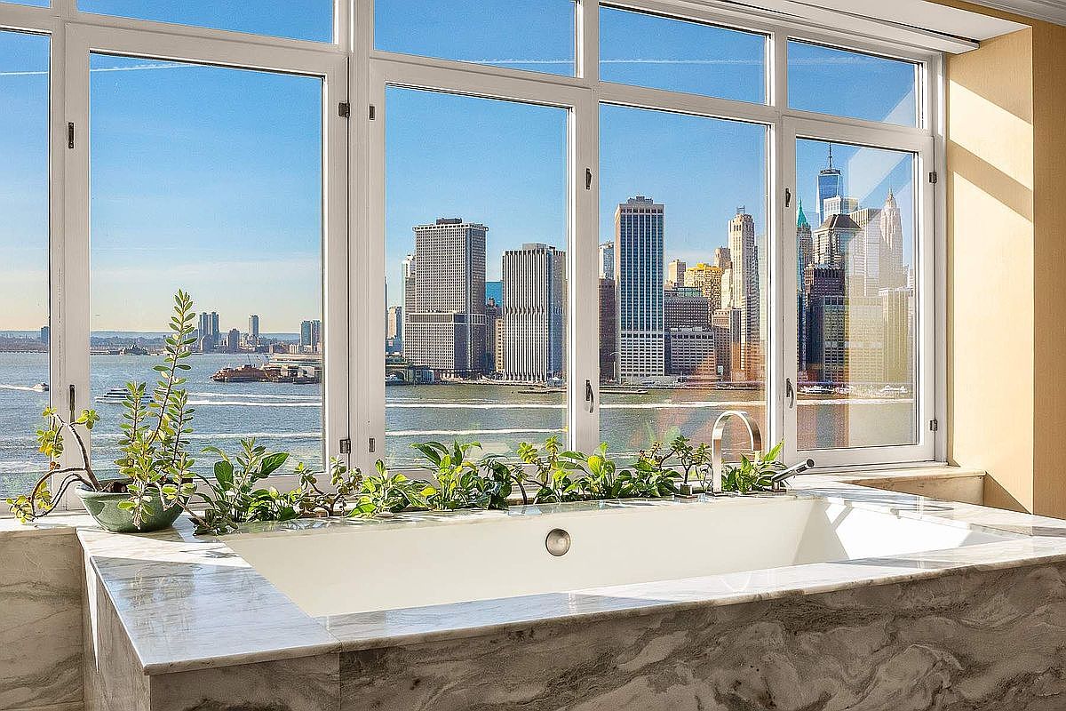 This luxurious primary bathroom features a deep soaking tub encased in elegant, veined marble, positioned directly against a floor-to-ceiling window wall. The expansive windows offer a breathtaking, panoramic view of the New York City skyline and the harbor, creating a serene and sophisticated atmosphere. Lush green plants line the ledge of the tub, adding a touch of natural warmth to the sleek, high-end interior design.