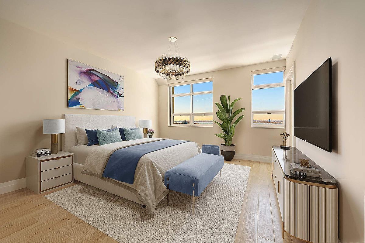 This bright and airy primary bedroom features a contemporary design with a neutral color palette accented by soft blue tones. The room is anchored by a plush bed with a tufted headboard, complemented by a matching bench, sleek nightstands, and a large wall-mounted television. Large windows provide ample natural light and a scenic view, while a potted plant and modern chandelier add a touch of elegance to the space.