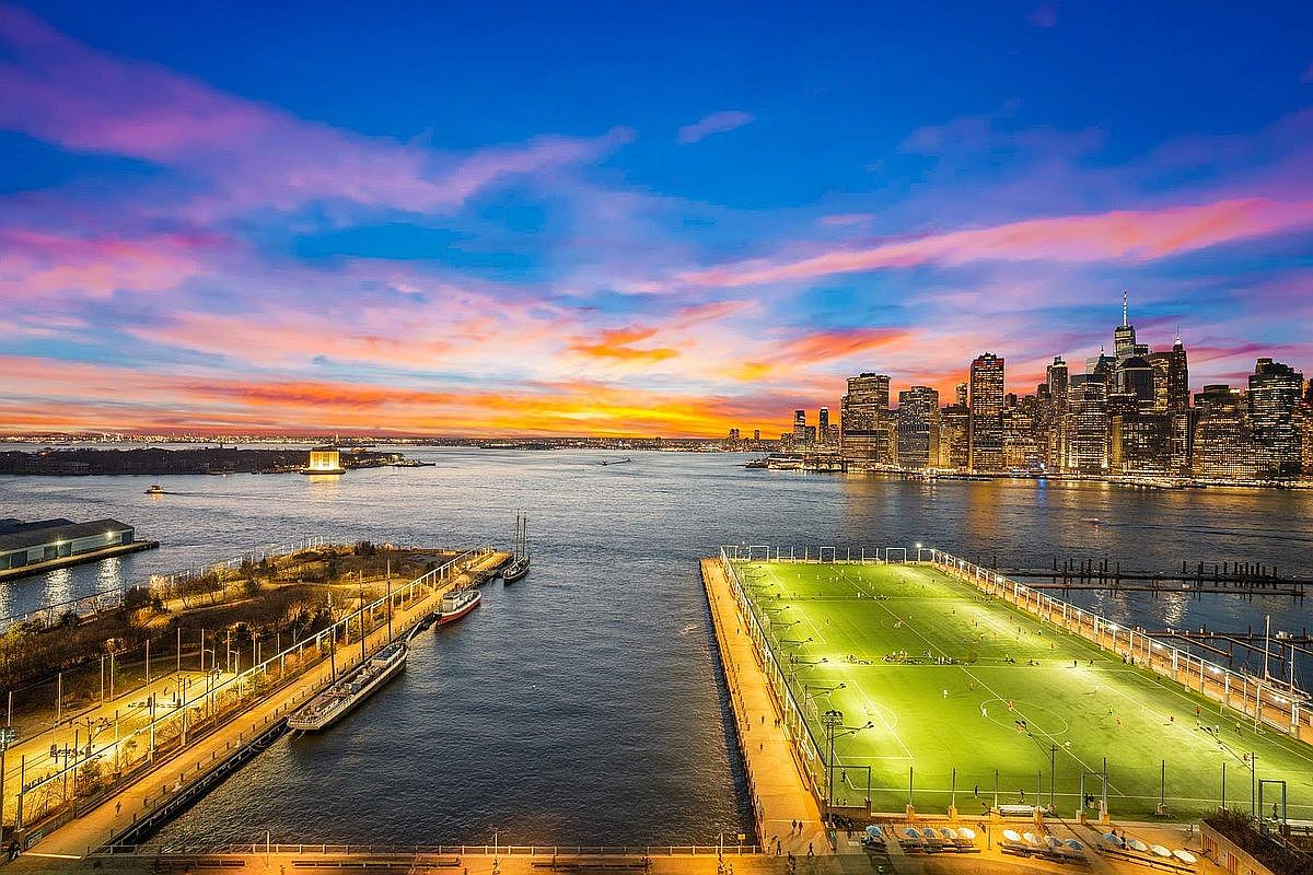 This stunning aerial view captures a vibrant sunset over the New York City skyline, highlighting the waterfront location of Brooklyn Bridge Park. The foreground features a brightly illuminated soccer field on a pier, contrasting with the calm harbor waters and the iconic Manhattan skyscrapers in the distance. The composition offers a cinematic perspective, emphasizing the prime urban lifestyle and recreational amenities available in this highly desirable waterfront community.