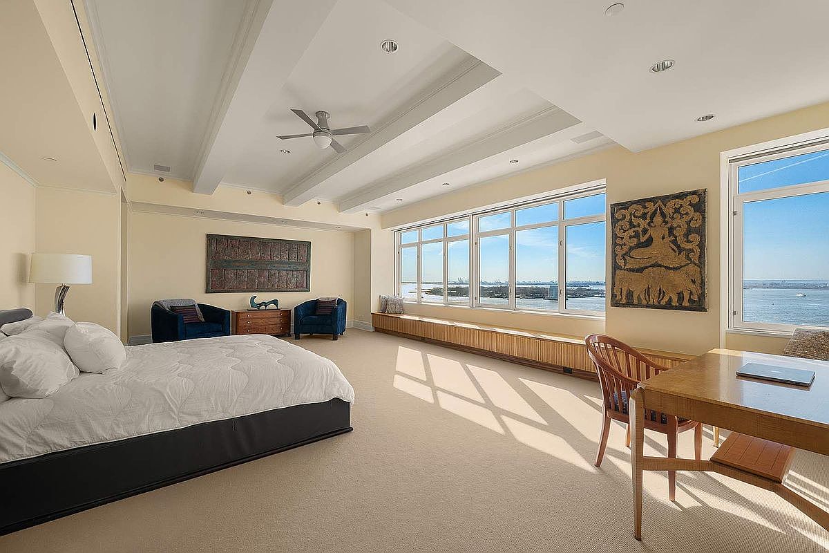This spacious primary bedroom features a neutral color palette, plush carpeting, and a large bed positioned to face expansive windows with panoramic water views. The room includes a dedicated workspace with a wooden desk and chair, as well as a cozy seating area with two blue armchairs. The high, tray-style ceiling with recessed lighting and a ceiling fan adds an elegant, airy feel to this bright and serene living space.