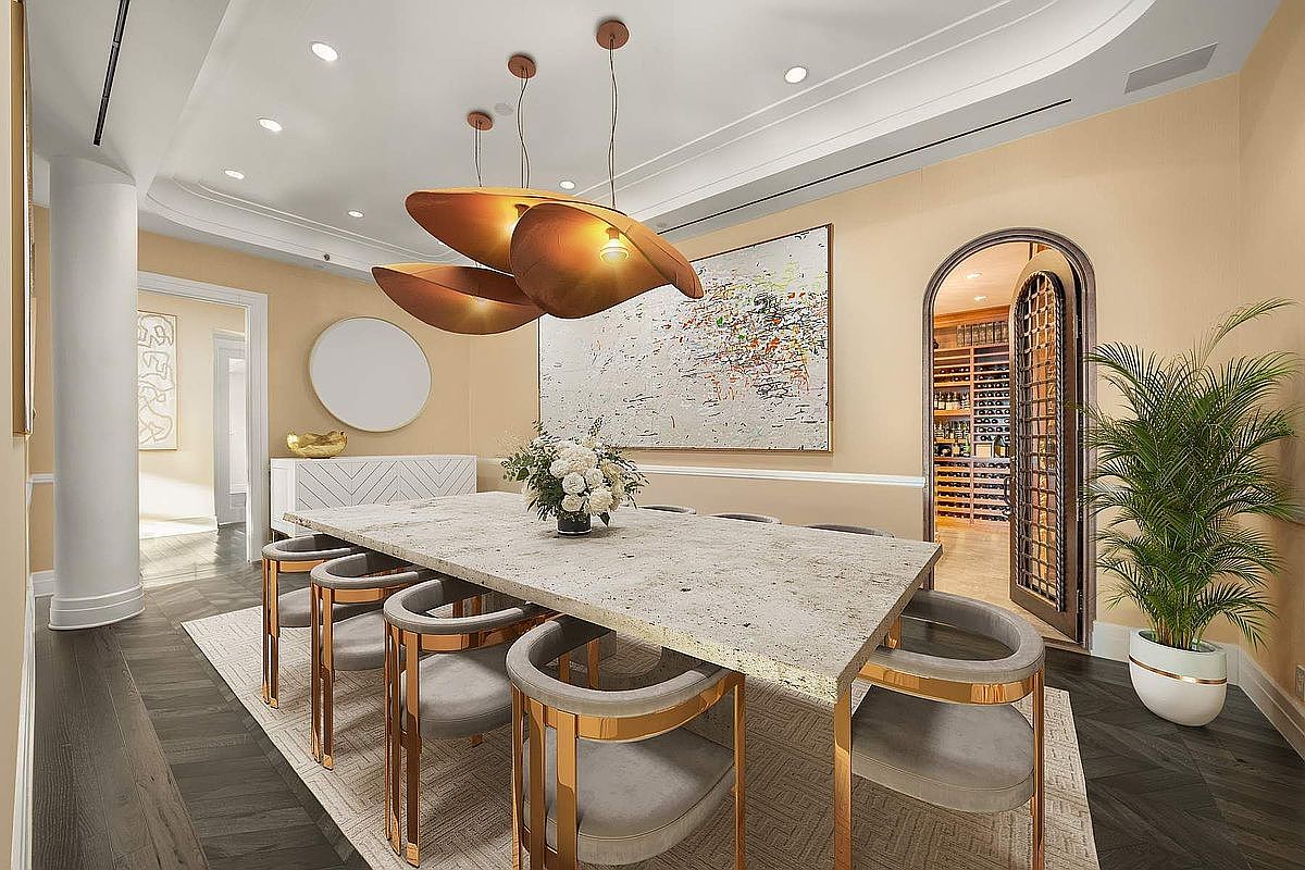 This elegant dining room features a large, light-colored stone table surrounded by modern, gold-accented velvet chairs, set beneath a striking sculptural pendant light fixture. The room is anchored by a textured area rug and a large abstract painting on the wall, while an arched doorway leads into a sophisticated, well-lit wine cellar. The overall aesthetic is one of refined luxury, characterized by warm neutral tones, high-end finishes, and a spacious, inviting atmosphere.