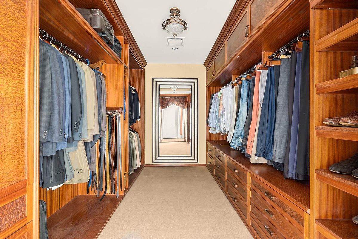 This spacious, high-end walk-in closet features rich, warm-toned custom wood cabinetry lining both sides of a central aisle. The space is organized with hanging rods for clothing, integrated drawers, and open shelving for accessories, all illuminated by a classic ceiling light fixture. A large, framed mirror at the far end adds depth to the room, creating a sophisticated and orderly atmosphere.