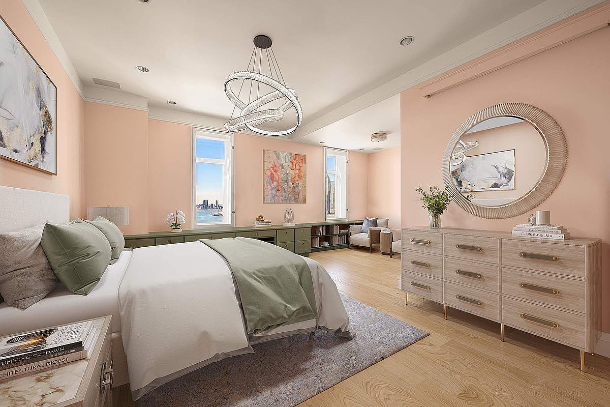 This elegant primary bedroom features a soft, calming color palette with blush pink walls and sage green accents. The room is anchored by a modern, sculptural chandelier and a light wood dresser topped with a large, textured circular mirror. A low-profile bed with plush bedding sits on a neutral area rug, while a long, built-in storage unit runs along the window wall, offering both functionality and a seamless view of the outdoors.