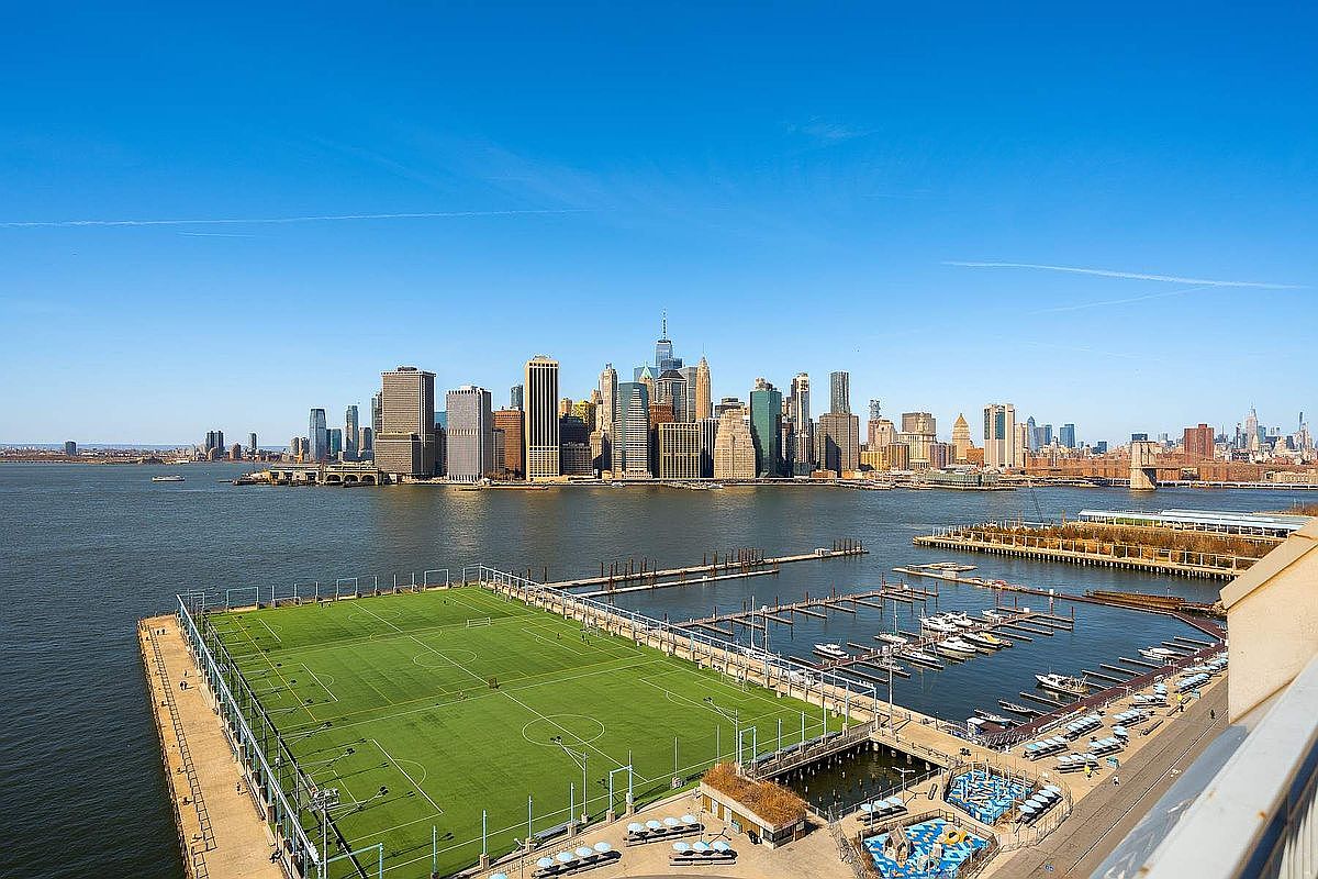 This high-angle aerial view captures a vibrant waterfront scene featuring a large, well-maintained soccer field situated on a pier. Adjacent to the field is a marina filled with boats, providing a dynamic contrast between recreational space and urban infrastructure. The background showcases a breathtaking, panoramic skyline of Manhattan, emphasizing the property's prime location and iconic city views.