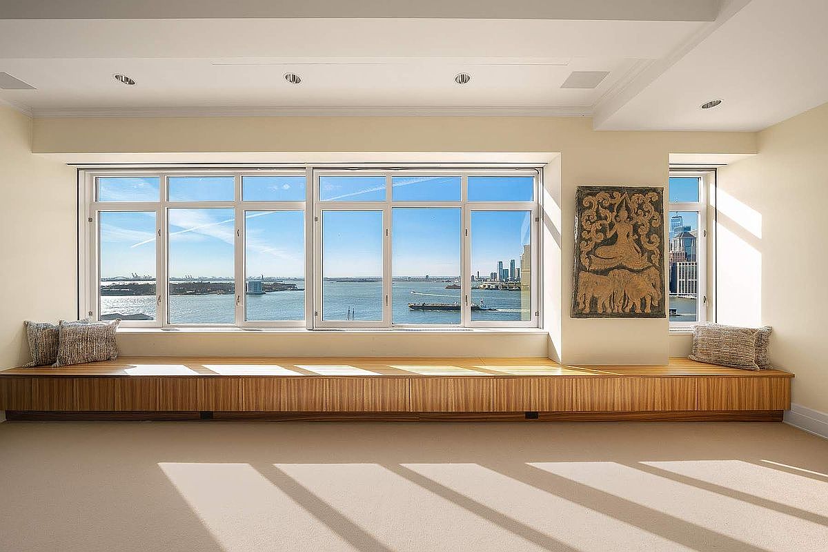 This bright and airy living space features a long, custom-built wooden window seat that spans the width of the room, offering panoramic waterfront views. The minimalist design is complemented by neutral carpeting and a large, textured wall art piece, creating a serene and sophisticated atmosphere. The perspective is head-on, emphasizing the expansive windows and the seamless integration of indoor comfort with the scenic outdoor landscape.