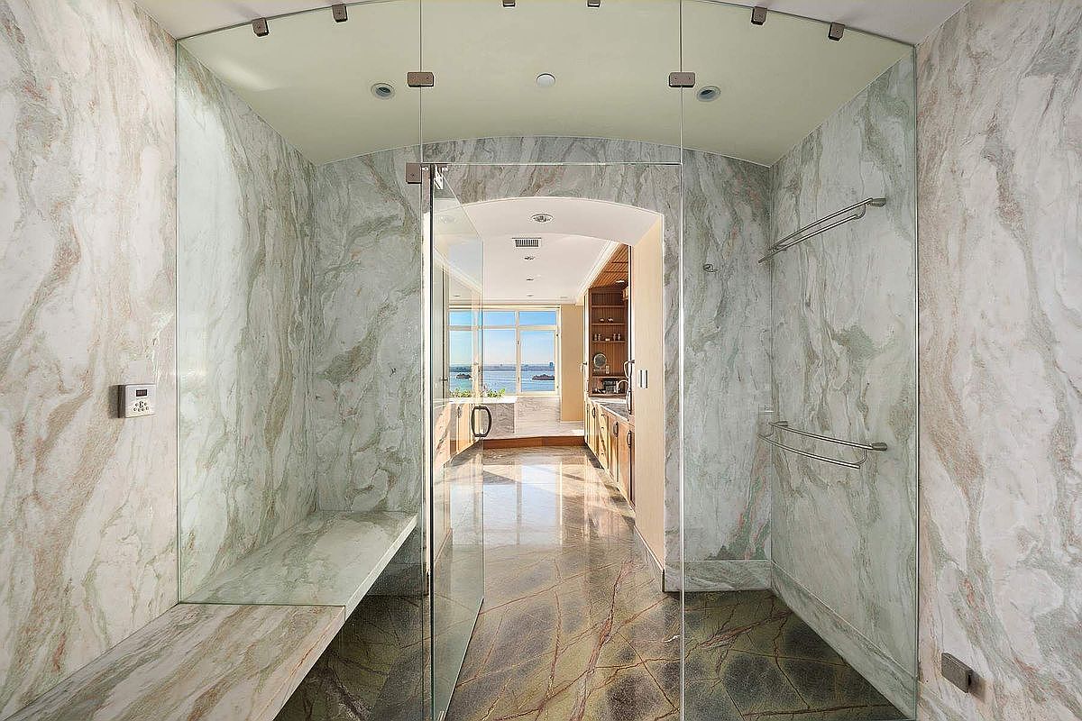 This luxurious, spa-inspired steam shower features floor-to-ceiling marble slab walls and a built-in stone bench, enclosed by seamless glass panels. The perspective looks through the shower into a bright, adjacent bathroom area with a view of the ocean through a large window. The space exudes high-end elegance with its polished stone surfaces and sophisticated, minimalist design.