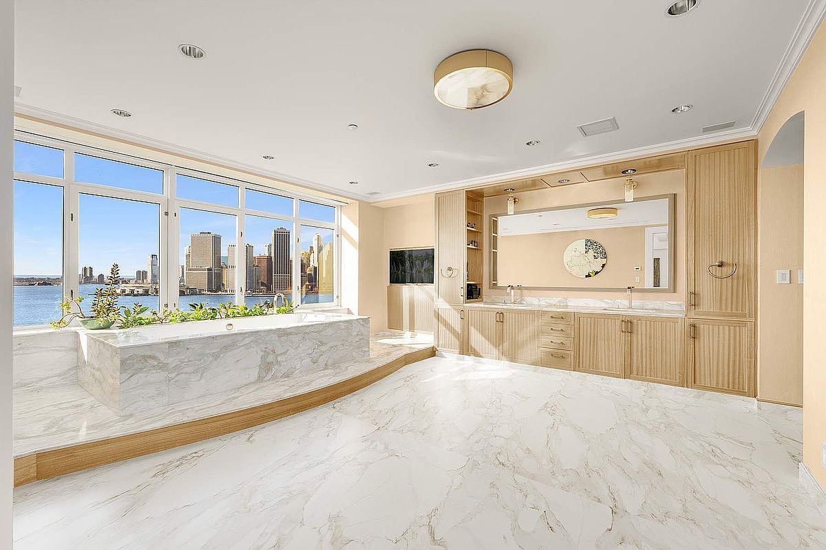 This luxurious primary bathroom features a stunning marble-clad soaking tub positioned directly in front of floor-to-ceiling windows that offer a breathtaking panoramic view of the city skyline and waterfront. The space is anchored by a custom light-wood vanity with dual sinks and a large, integrated mirror, all set against elegant marble flooring. The overall aesthetic is one of sophisticated, high-end urban living, blending natural materials with a dramatic, open-concept design.