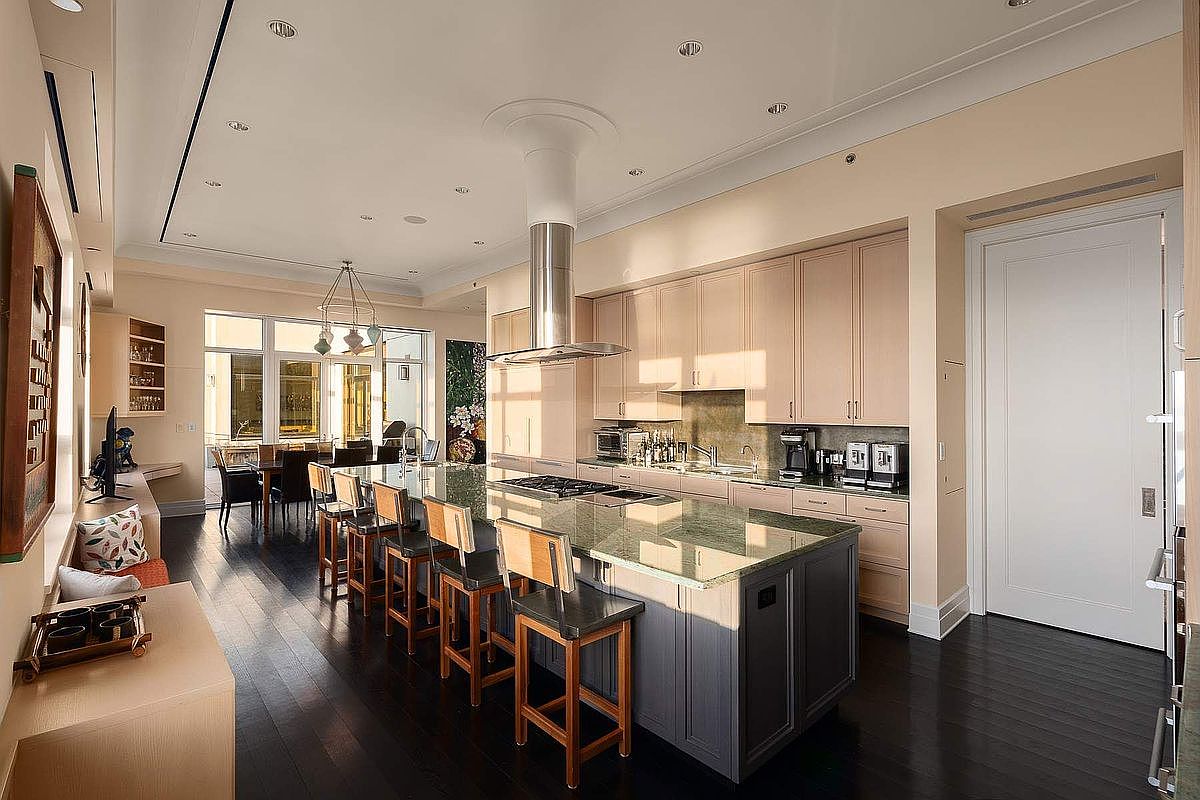 This spacious, high-end kitchen features a large central island with a dark base and stone countertop, complemented by light-toned cabinetry and dark hardwood flooring. A sleek stainless steel range hood hangs above the island, while recessed lighting and a nearby dining area create an open, airy atmosphere. The perspective captures the depth of the room, showcasing a seamless blend of modern functionality and elegant design.