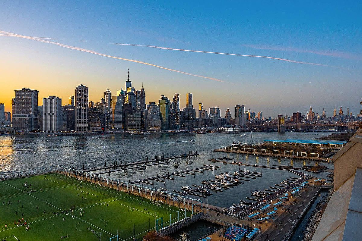 This stunning aerial perspective captures a panoramic view of the New York City skyline at dusk, framed by the East River. In the foreground, a vibrant green soccer field and a bustling marina with docked boats provide a dynamic contrast to the towering skyscrapers across the water. The golden hour light casts a warm glow over the urban landscape, creating an inviting and sophisticated atmosphere.