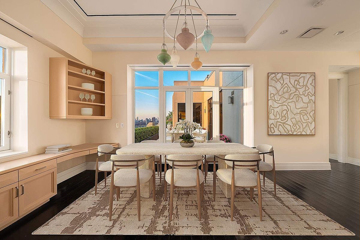 This elegant dining room features a sophisticated, neutral color palette with a textured area rug anchoring a modern stone-topped table surrounded by six upholstered chairs. A unique, multi-colored glass pendant chandelier serves as a focal point above the table, while built-in light wood cabinetry and a large abstract art piece add depth to the space. The room opens through floor-to-ceiling glass doors to a terrace, offering a bright, airy atmosphere with a view of the city skyline.