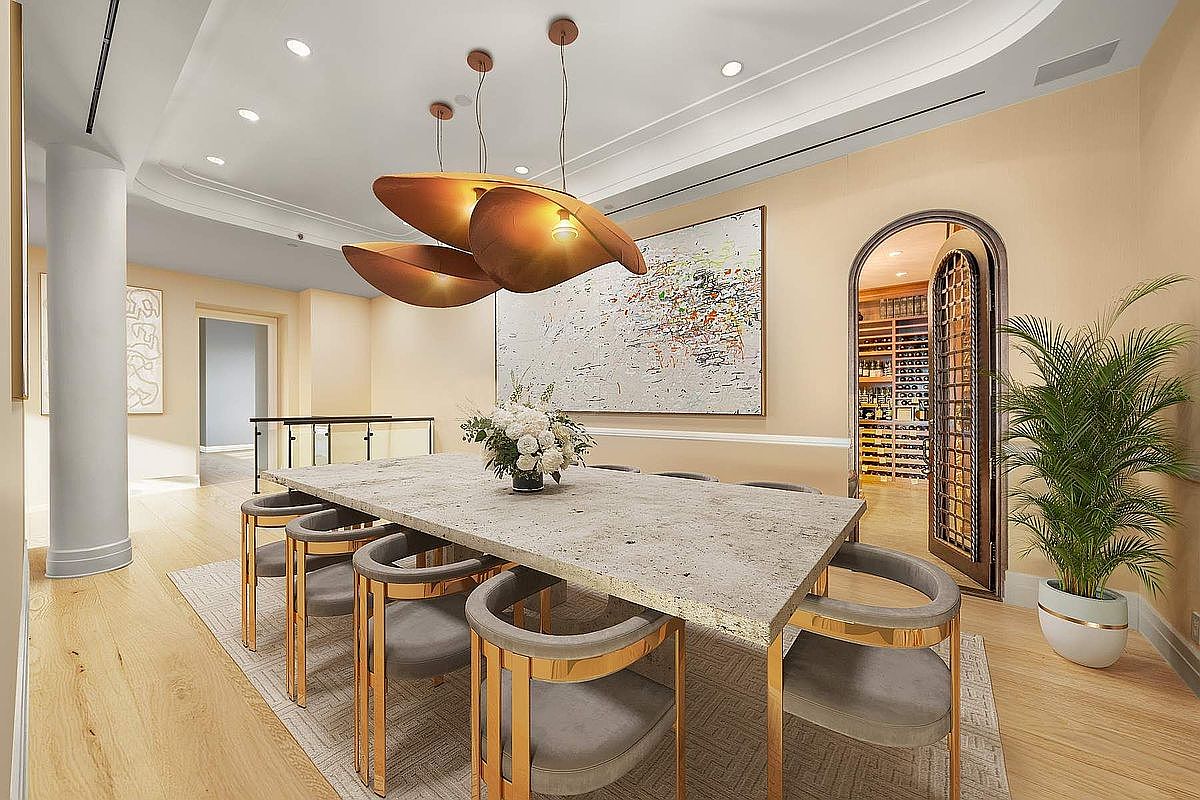 This elegant dining room features a large, textured stone-top table surrounded by modern, gold-accented velvet chairs, centered beneath a striking sculptural pendant light fixture. The space is anchored by a large abstract painting on the wall and a sophisticated arched wine cellar entrance, creating a luxurious and refined atmosphere. The perspective is a wide-angle shot that captures the seamless flow between the dining area, the wine cellar, and the adjacent hallway.