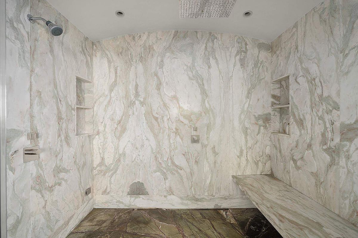 This luxurious walk-in shower features stunning, book-matched marble wall slabs with intricate veining and a matching stone bench. The space is equipped with a rainfall showerhead, a wall-mounted showerhead, and recessed shelving niches, all set against a dark, textured stone floor. The perspective is a centered, eye-level shot that emphasizes the high-end materials and spacious, spa-like design.