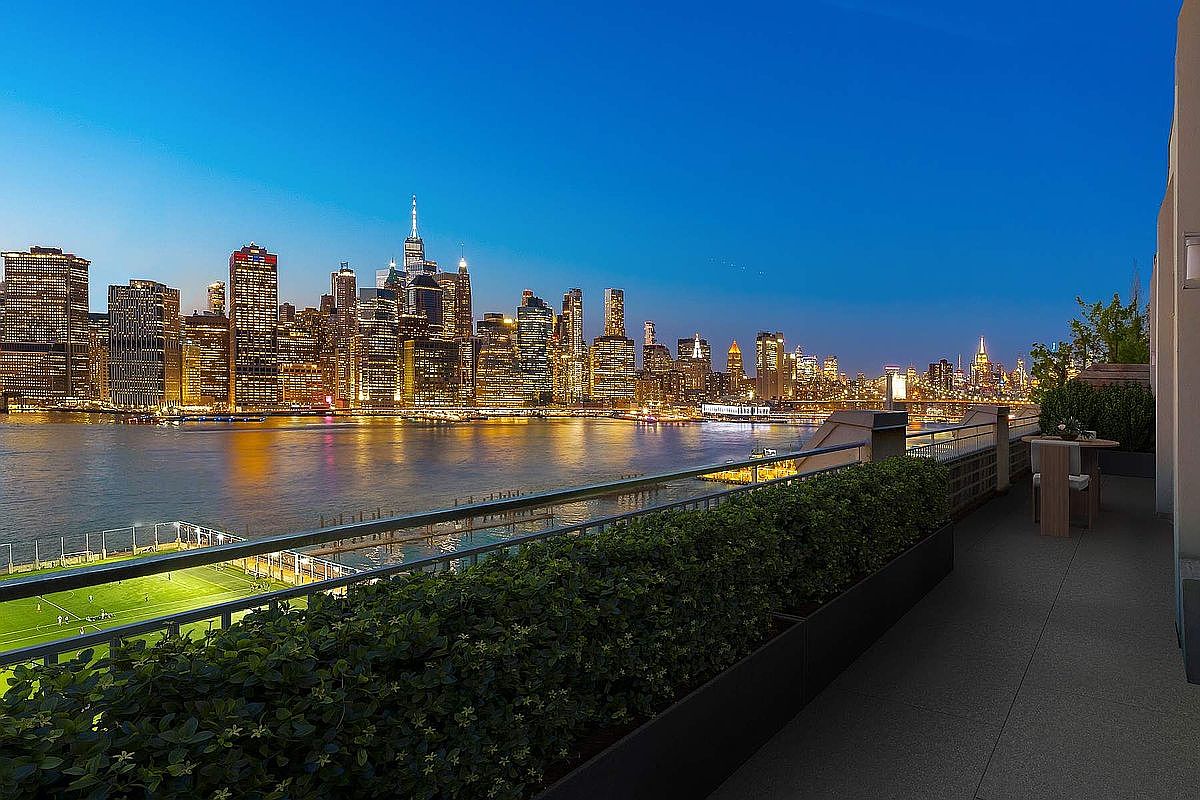 This stunning balcony offers a breathtaking, unobstructed panoramic view of the New York City skyline at dusk, with the city lights beginning to glow against the deep blue sky. The space features a sleek metal railing, a manicured hedge planter, and a small bistro table, creating an intimate and sophisticated outdoor living area. The perspective captures the dramatic contrast between the lush greenery of the balcony and the expansive urban landscape across the water.