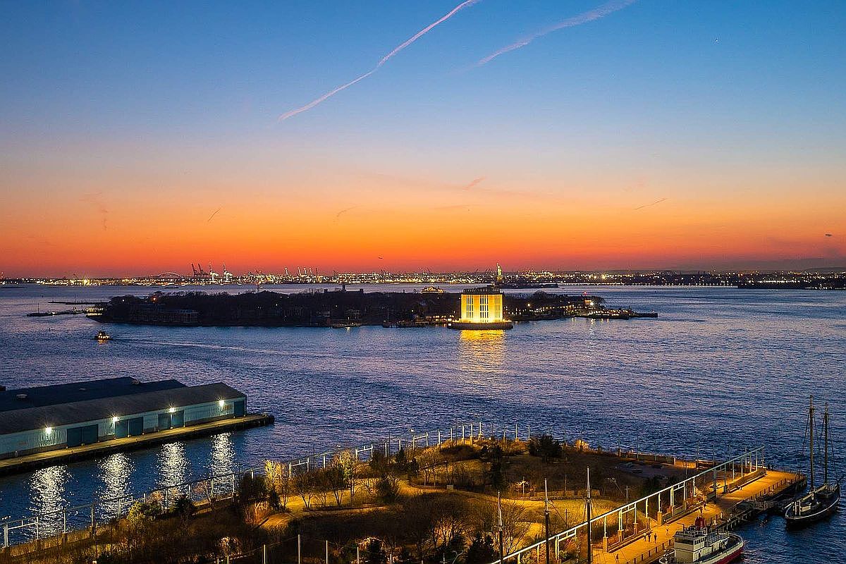 This stunning aerial view captures a serene sunset over New York Harbor, featuring the illuminated Castle Clinton National Monument on the island in the foreground. The composition highlights the juxtaposition of the tranquil water, the historic structure, and the distant city skyline glowing under a vibrant orange and blue sky. The perspective provides a cinematic sense of scale, emphasizing the prime waterfront location and the peaceful evening atmosphere.