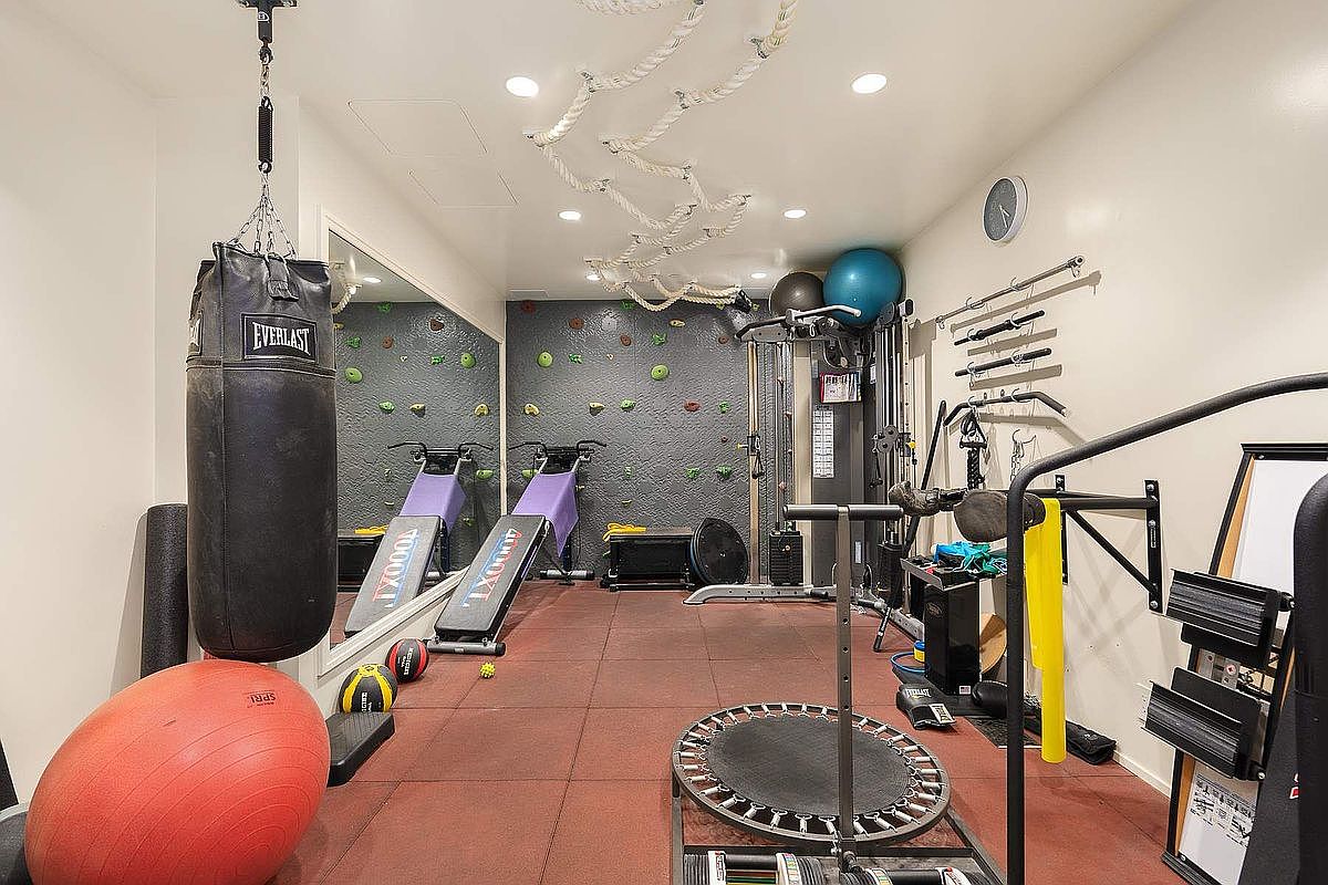 This well-equipped home gym features a dedicated climbing wall, a heavy punching bag, and multiple exercise machines including incline benches and a mini trampoline. The space is finished with durable rubber flooring, recessed lighting, and a large wall mirror that enhances the room's depth. It offers a comprehensive and versatile workout environment designed for a variety of fitness activities.