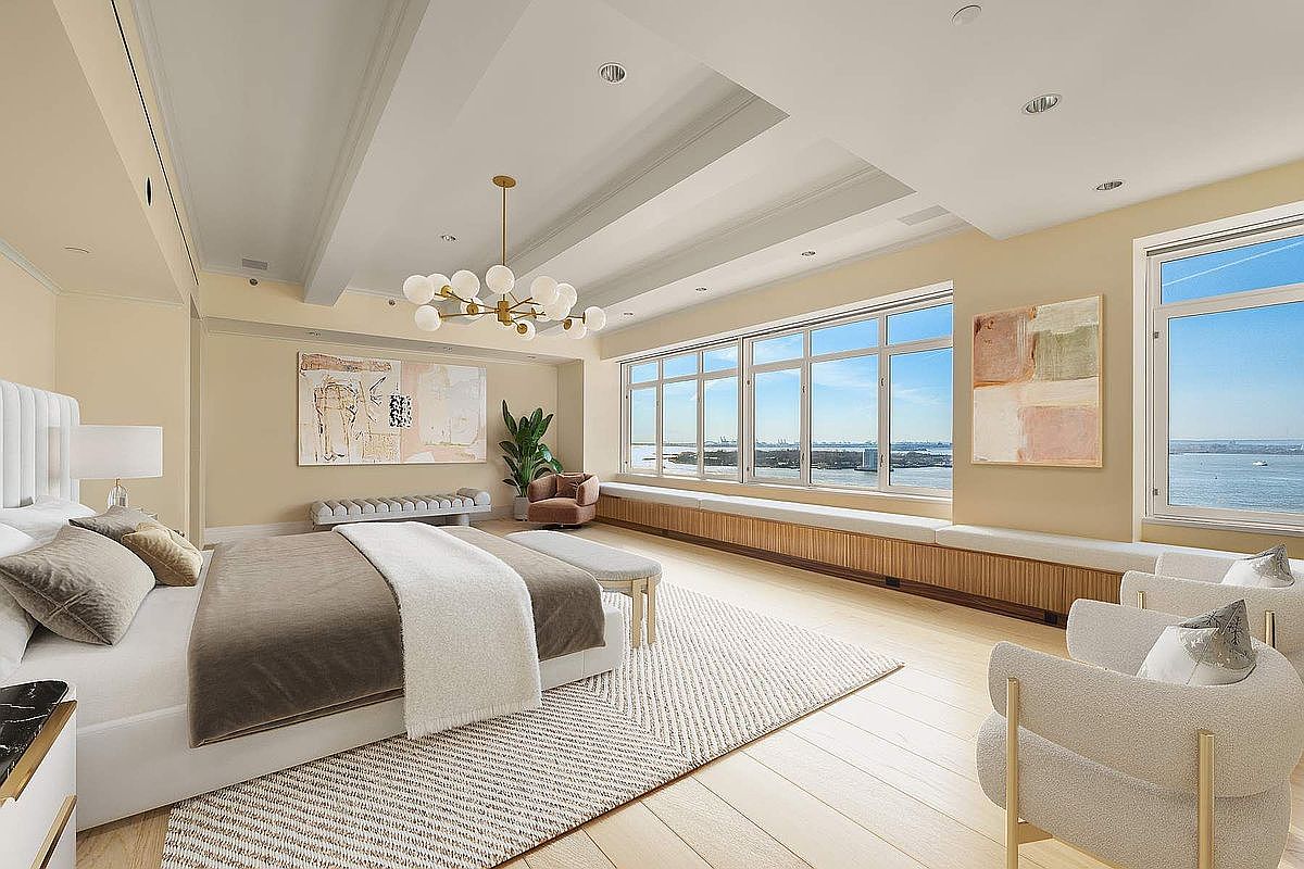 This luxurious primary bedroom features a sophisticated, neutral color palette and expansive floor-to-ceiling windows that offer breathtaking panoramic water views. The room is anchored by a plush bed with high-end linens, complemented by elegant modern armchairs and a custom built-in window seat. A striking contemporary chandelier serves as a focal point, enhancing the airy, high-ceilinged atmosphere of this serene waterfront retreat.