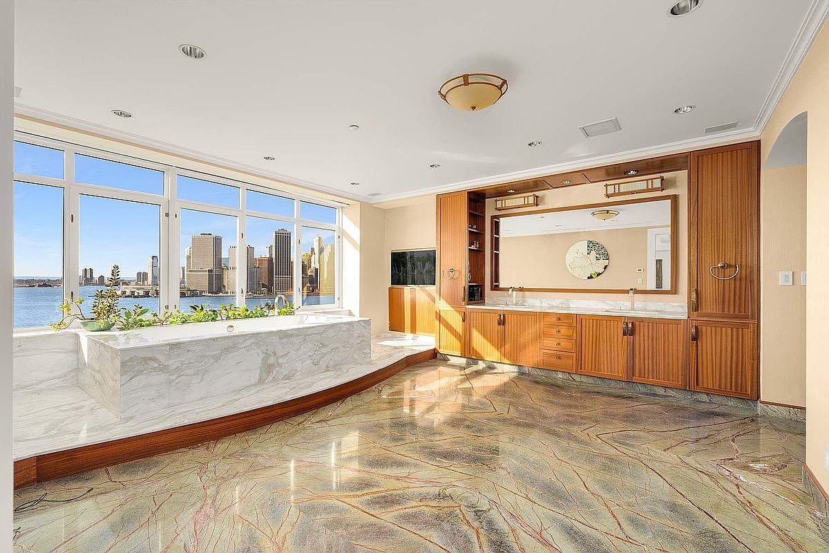This luxurious primary bathroom features a stunning marble-clad soaking tub positioned directly in front of floor-to-ceiling windows that offer a breathtaking panoramic view of the city skyline and waterfront. The room is anchored by a polished, veined marble floor and custom wood cabinetry with a large integrated vanity mirror. The overall aesthetic is one of high-end sophistication, blending natural stone textures with warm wood tones to create a serene, spa-like atmosphere.