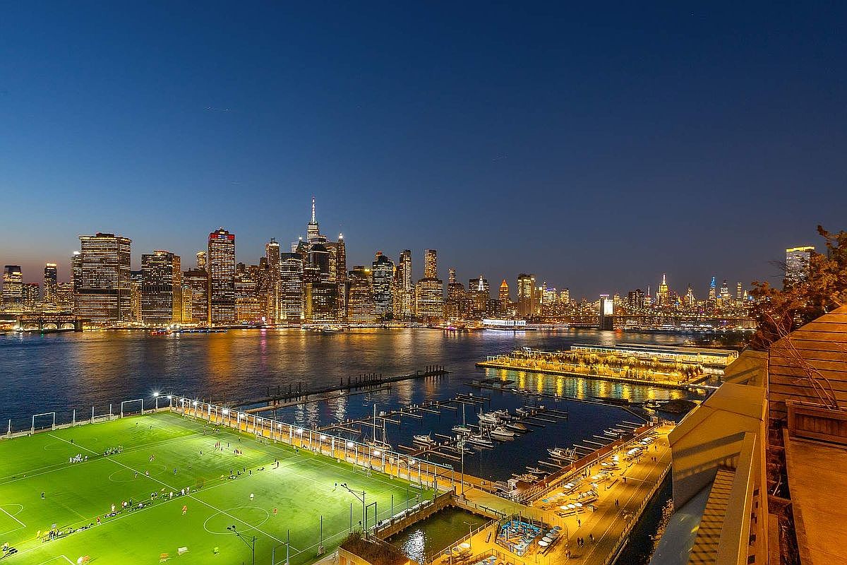This high-angle shot captures a stunning, panoramic view of the Manhattan skyline at dusk from a private balcony. The foreground features a vibrant, illuminated soccer field and a marina, creating a dynamic contrast between urban activity and the tranquil waterfront. The composition emphasizes the luxury of the location, showcasing a prime vantage point for enjoying the city's iconic evening lights.