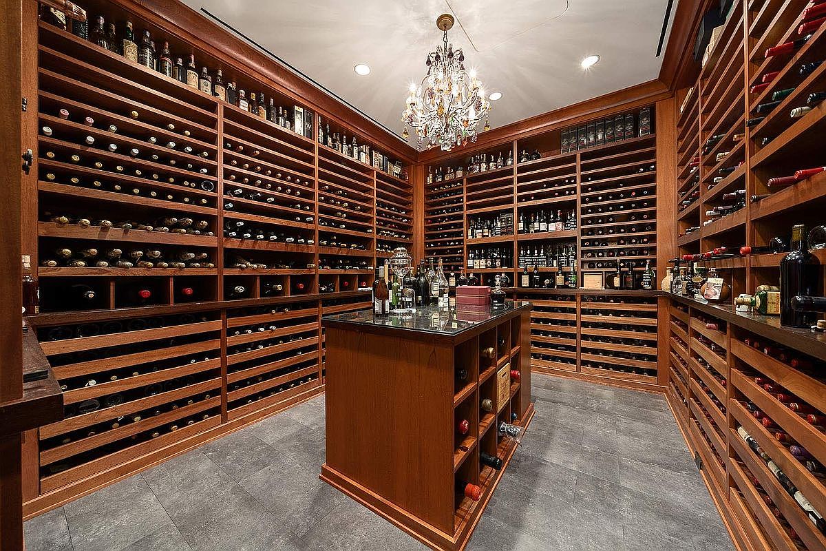 This sophisticated, custom-built wine cellar features extensive floor-to-ceiling wooden racking systems designed to house a large collection of wine bottles. A central island with a dark stone countertop provides a focal point, while an elegant crystal chandelier hangs from the ceiling, adding a touch of luxury to the space. The room is finished with dark grey tile flooring, creating a refined and temperature-controlled environment perfect for wine enthusiasts.