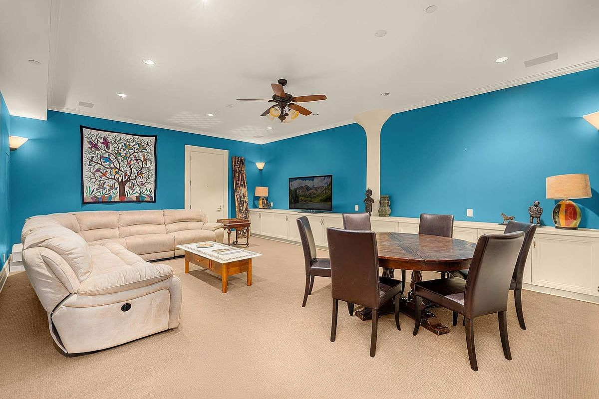 This spacious, open-concept living and dining area features vibrant blue walls and neutral carpeting, creating a bold yet comfortable atmosphere. The room is furnished with a large, plush sectional sofa and a circular dining table, making it ideal for both relaxation and entertaining. A ceiling fan, recessed lighting, and built-in cabinetry provide functional and aesthetic appeal to this versatile living space.