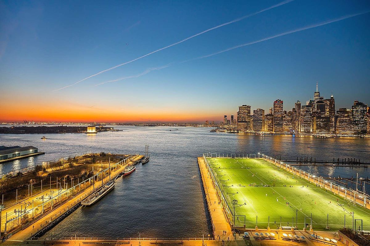 This stunning aerial view captures a vibrant waterfront scene at dusk, featuring a brightly illuminated soccer field on a pier in the foreground. The composition offers a panoramic perspective of the New York City skyline across the water, set against a beautiful sunset gradient. The juxtaposition of the active, glowing recreational space and the iconic urban backdrop creates a sophisticated and highly desirable metropolitan atmosphere.