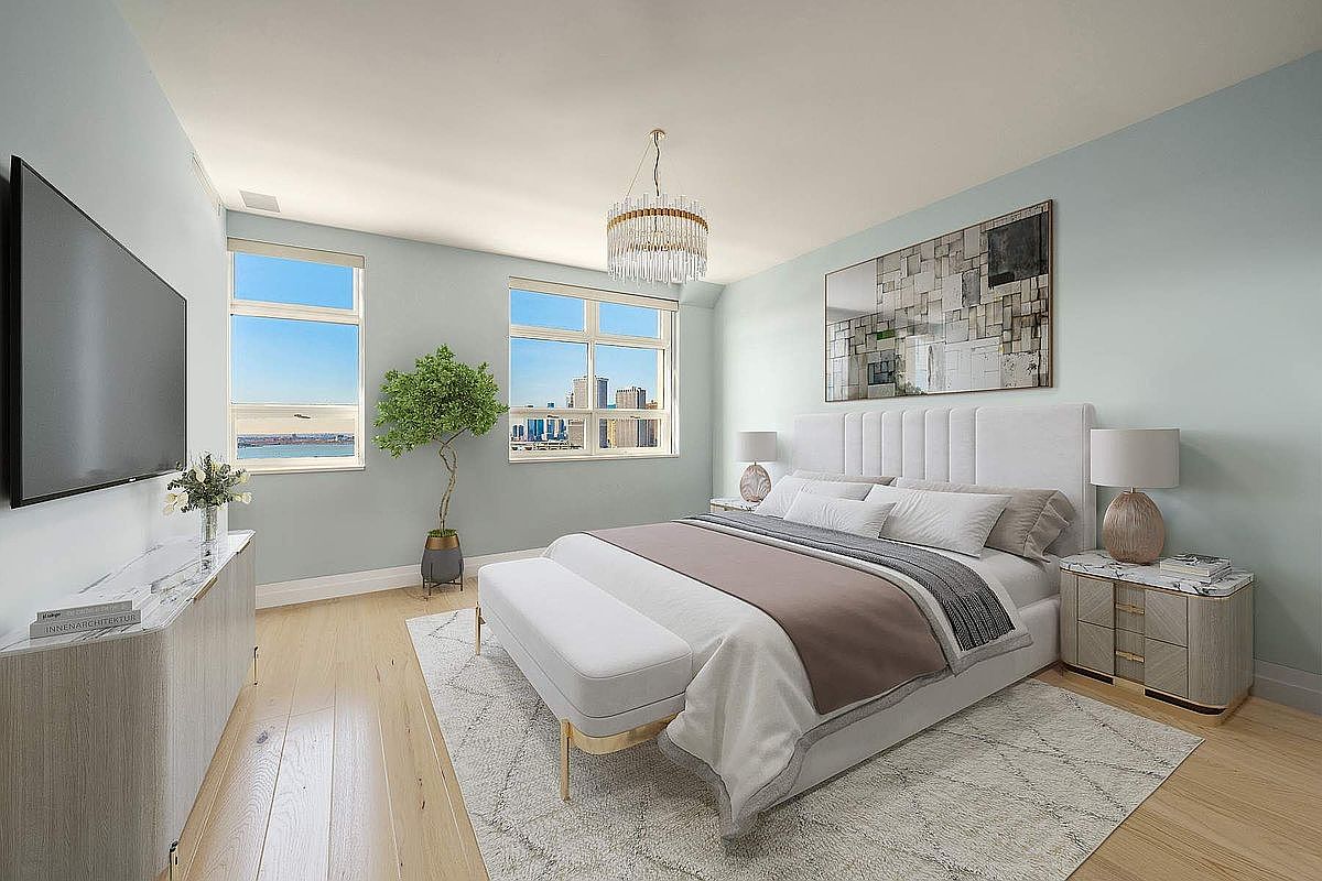 This elegant primary bedroom features a sophisticated, neutral color palette with soft blue walls and light hardwood flooring. The room is anchored by a plush, upholstered bed with a tall headboard, flanked by matching nightstands and lamps, while a crystal chandelier adds a touch of luxury. Large windows provide ample natural light and a scenic view, creating a serene and airy atmosphere perfect for a high-end residence.