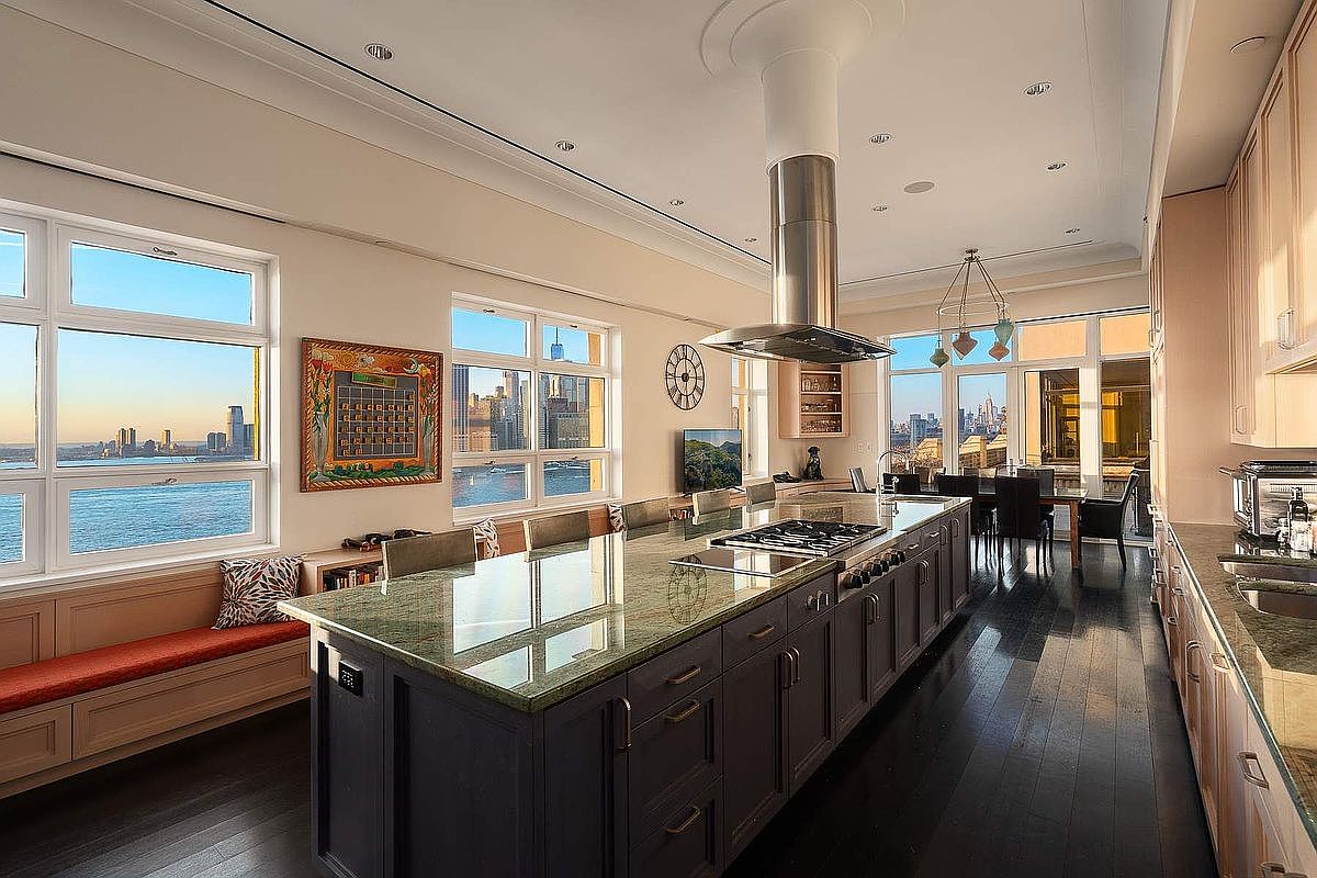 This expansive, high-end kitchen features a massive central island with a dark base and a polished green granite countertop, serving as the focal point of the room. The space is bathed in natural light from large windows that offer panoramic city and waterfront views, complemented by dark hardwood flooring and sleek, modern cabinetry. A stainless steel range hood hangs above the island, while a built-in window seat provides a cozy spot to enjoy the impressive urban backdrop.