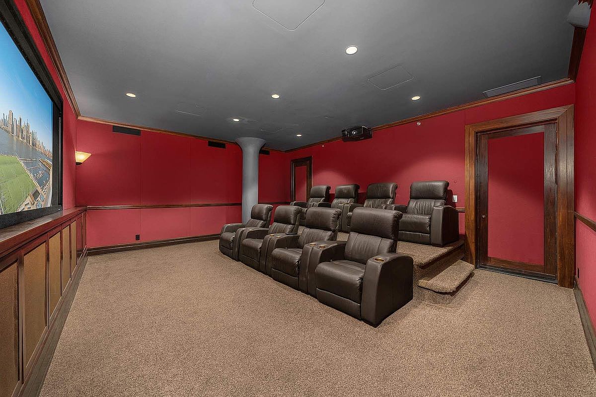 This dedicated home theater room features tiered seating with plush, dark leather recliners arranged in two rows. The space is defined by bold red walls, a dark grey ceiling, and a large projection screen, creating an immersive cinematic environment. A structural column and wood-trimmed accents add architectural character to this private entertainment space.