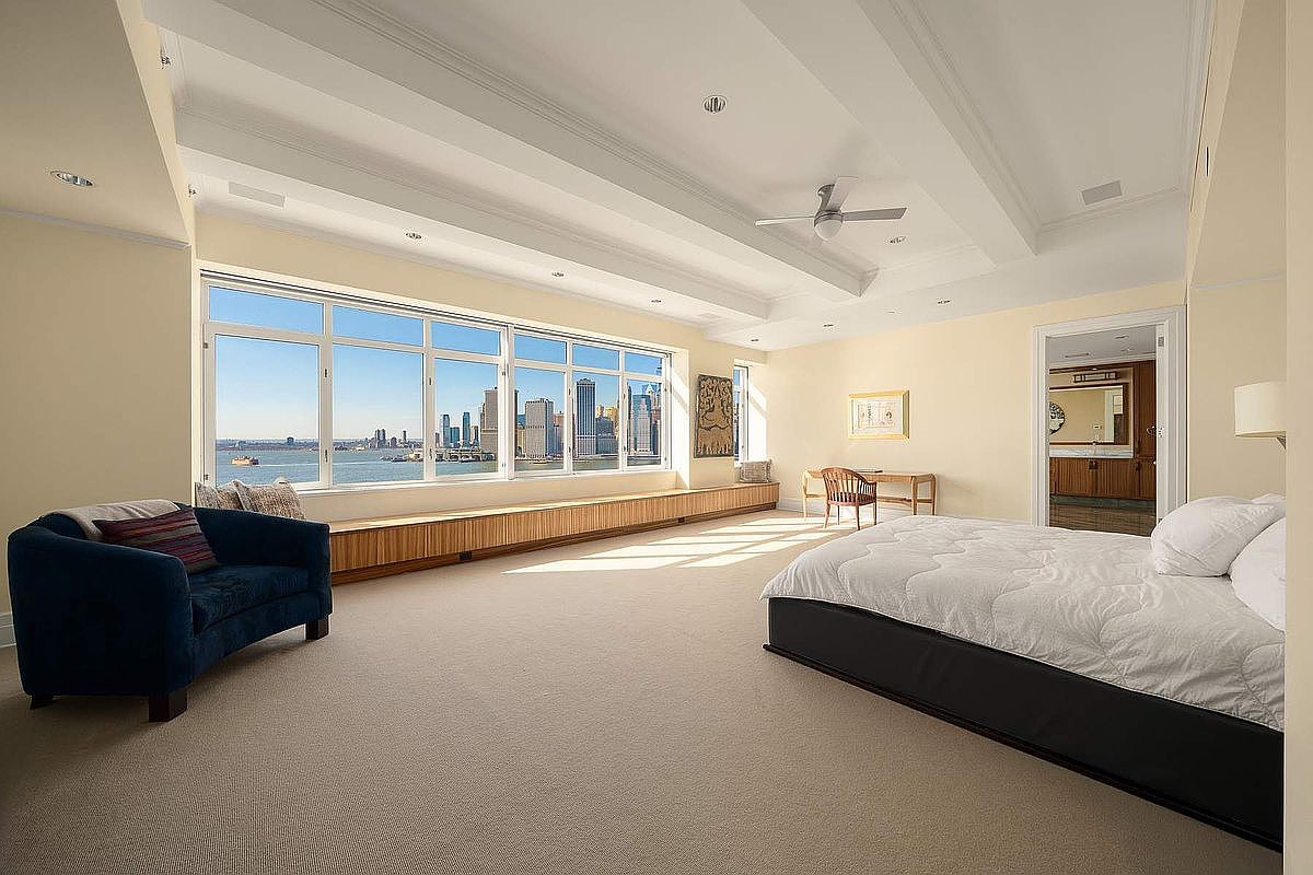 This expansive primary bedroom features a neutral color palette, plush carpeting, and a striking coffered ceiling with a central fan. A large wall of windows provides a panoramic view of the city skyline and waterfront, complemented by a long, built-in wooden bench. The room is furnished with a dark-framed bed and a cozy blue armchair, creating a serene and sophisticated atmosphere.