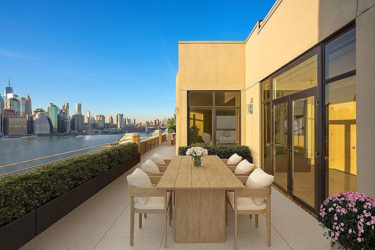 This luxurious, expansive terrace offers a sophisticated outdoor dining experience with a sleek wooden table and matching chairs set against a stunning panoramic view of the New York City skyline. The space is framed by manicured hedges and modern glass-paneled doors, creating a seamless transition between indoor and outdoor living. The bright, clear daylight highlights the clean architectural lines and the premium, high-end aesthetic of this urban oasis.