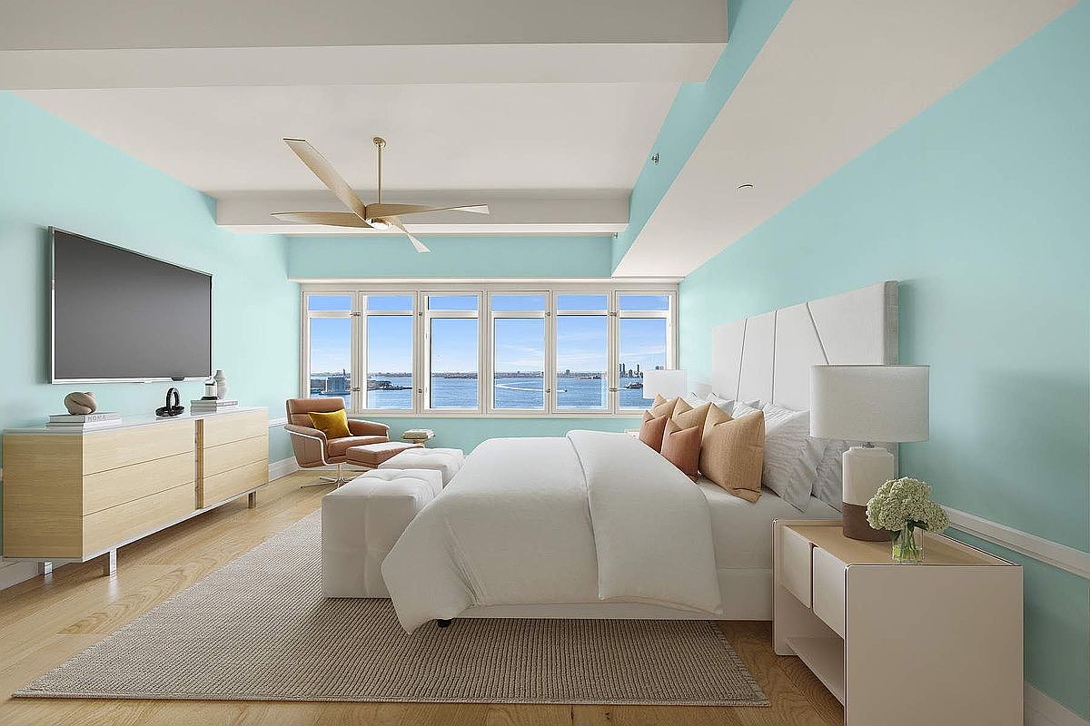 This bright and airy primary bedroom features a calming light blue color palette, a plush white bed, and a large window wall offering expansive water views. The room is furnished with a modern light-wood dresser, a sleek bedside table, and a stylish lounge chair, all set atop a textured area rug. The overall aesthetic is serene, sophisticated, and coastal-inspired, emphasizing natural light and a spacious, open layout.