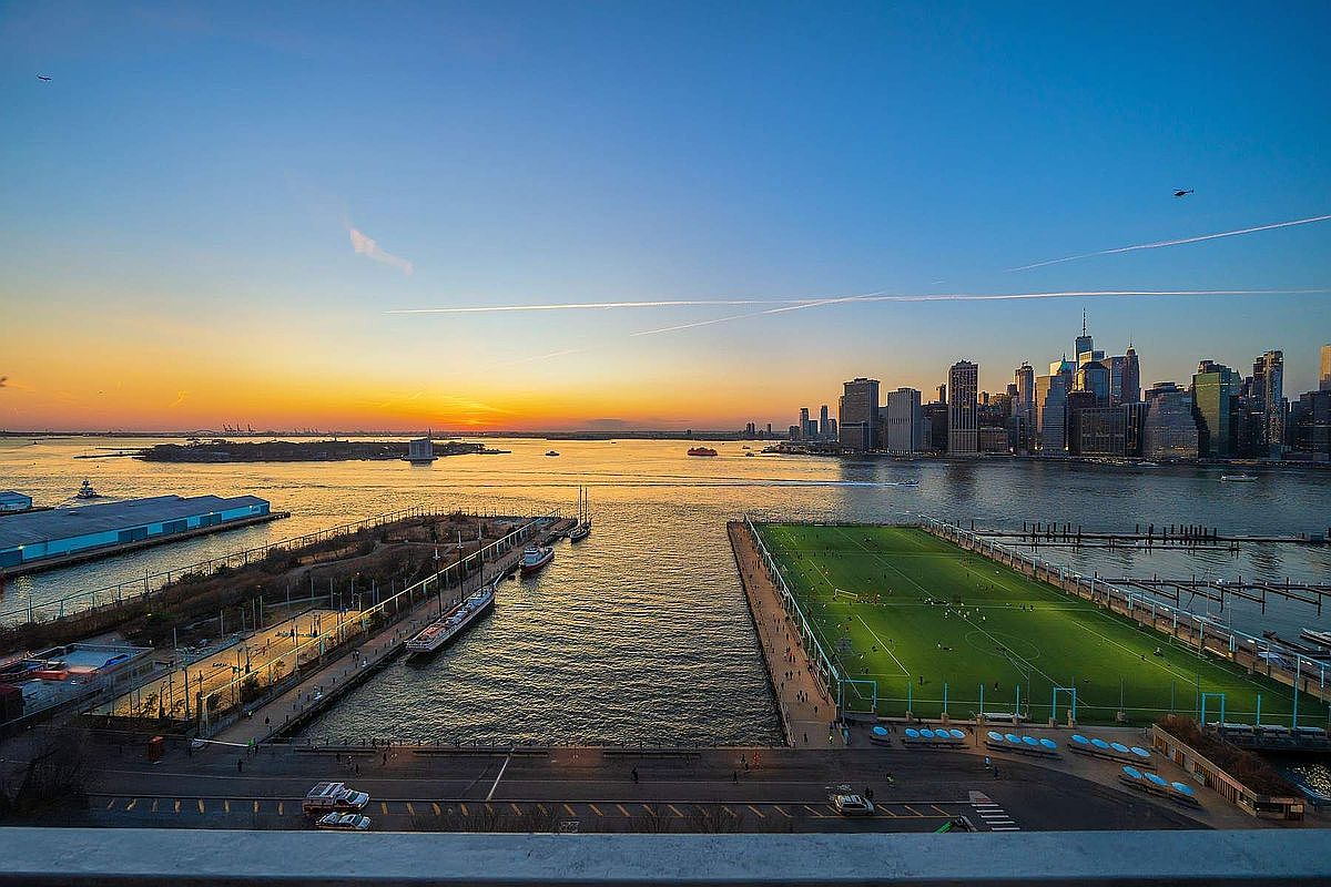 This stunning aerial view captures a vibrant waterfront scene at sunset, featuring a prominent soccer field on a pier and a bustling harbor. The composition highlights the dramatic contrast between the glowing orange horizon and the iconic New York City skyline in the distance. The perspective provides a cinematic sense of scale, emphasizing the property's prime location and proximity to major urban landmarks.