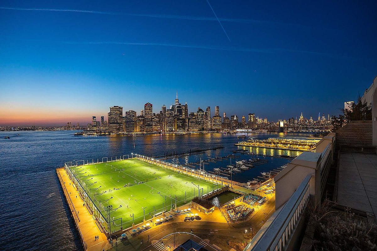 This stunning aerial view captures a vibrant waterfront scene at dusk, featuring a brightly illuminated soccer field situated on a pier extending into the river. In the background, the iconic New York City skyline glows with city lights against a deep blue twilight sky. The perspective is from a high-rise balcony, emphasizing the property's prime location and proximity to both recreational amenities and the urban center.