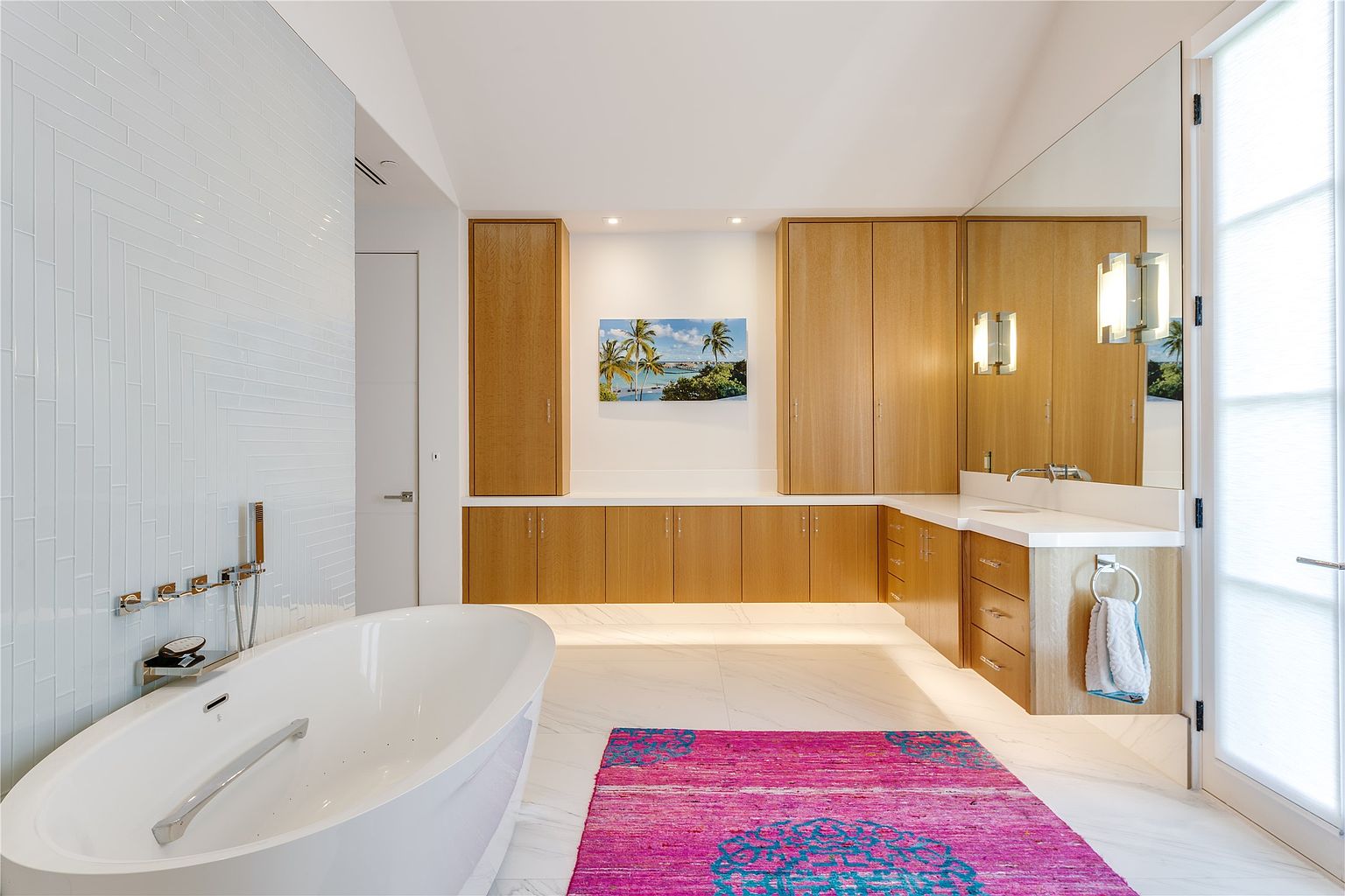 This bright and modern primary bathroom features a freestanding soaking tub set against a wall with light-colored, vertically stacked tile. A long vanity with light wood cabinetry and a white countertop extends along the wall, complemented by a large mirror and integrated lighting. A vibrant, patterned rug adds a pop of color to the white stone flooring, while a glass door provides access to the outdoors.