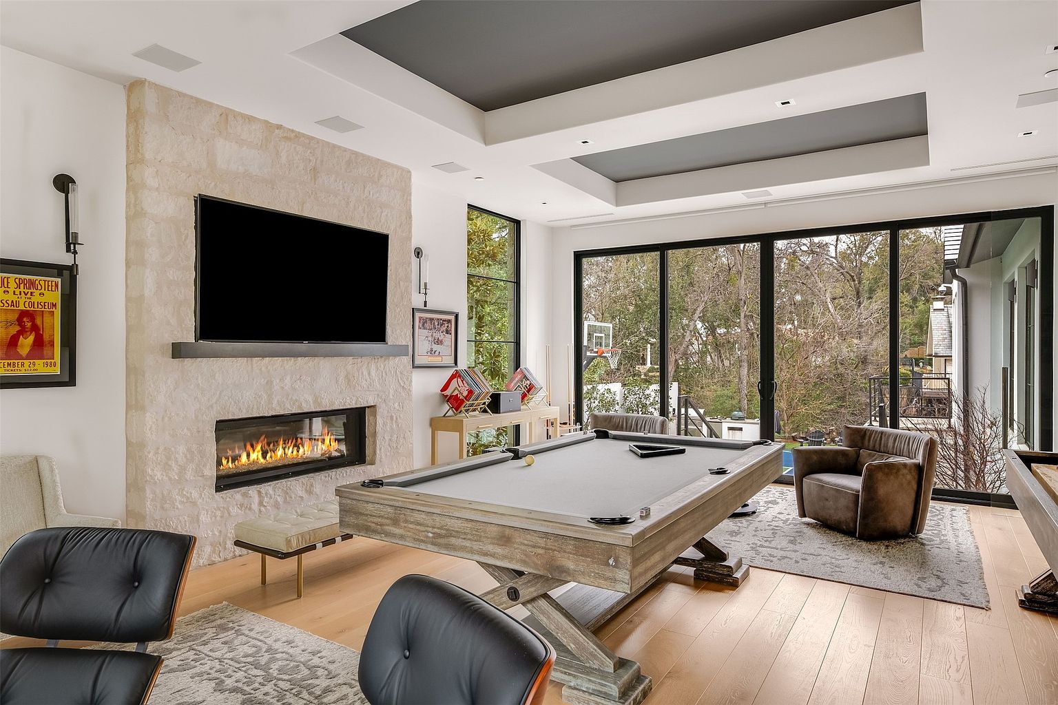 This spacious game room features a prominent pool table as the central focal point, set against a light wood floor and a stone-clad chimney breast with an integrated fireplace. Large glass sliding doors provide an expansive view of the wooded backyard, while a recessed ceiling detail adds architectural interest to the room. The space is furnished with comfortable leather armchairs and mid-century modern seating, creating a sophisticated yet relaxed atmosphere for entertainment.