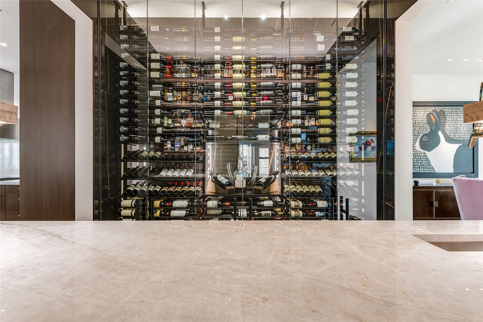 This sophisticated wine cellar features a glass-enclosed display wall with extensive horizontal bottle storage racks. In the foreground, a smooth, light-toned stone countertop provides a clean surface, while a framed piece of art depicting a rabbit with mathematical equations hangs on the adjacent wall. The perspective is centered, highlighting the organized collection and the modern, high-end aesthetic of the space.