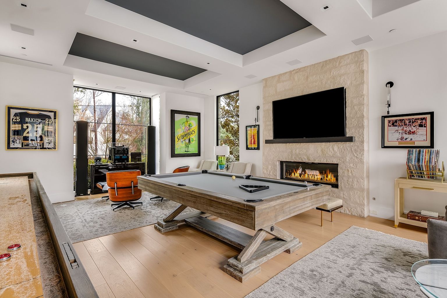 This sophisticated game room features a central pool table with a gray felt surface, positioned in front of a stone-clad chimney breast housing a linear fireplace. The space is accented by a dark-toned recessed ceiling, large windows that provide ample natural light, and a mix of modern and classic decor, including framed sports memorabilia and a Schweppes advertisement. The room is finished with light wood flooring and a textured area rug, creating a refined yet relaxed atmosphere for entertainment.