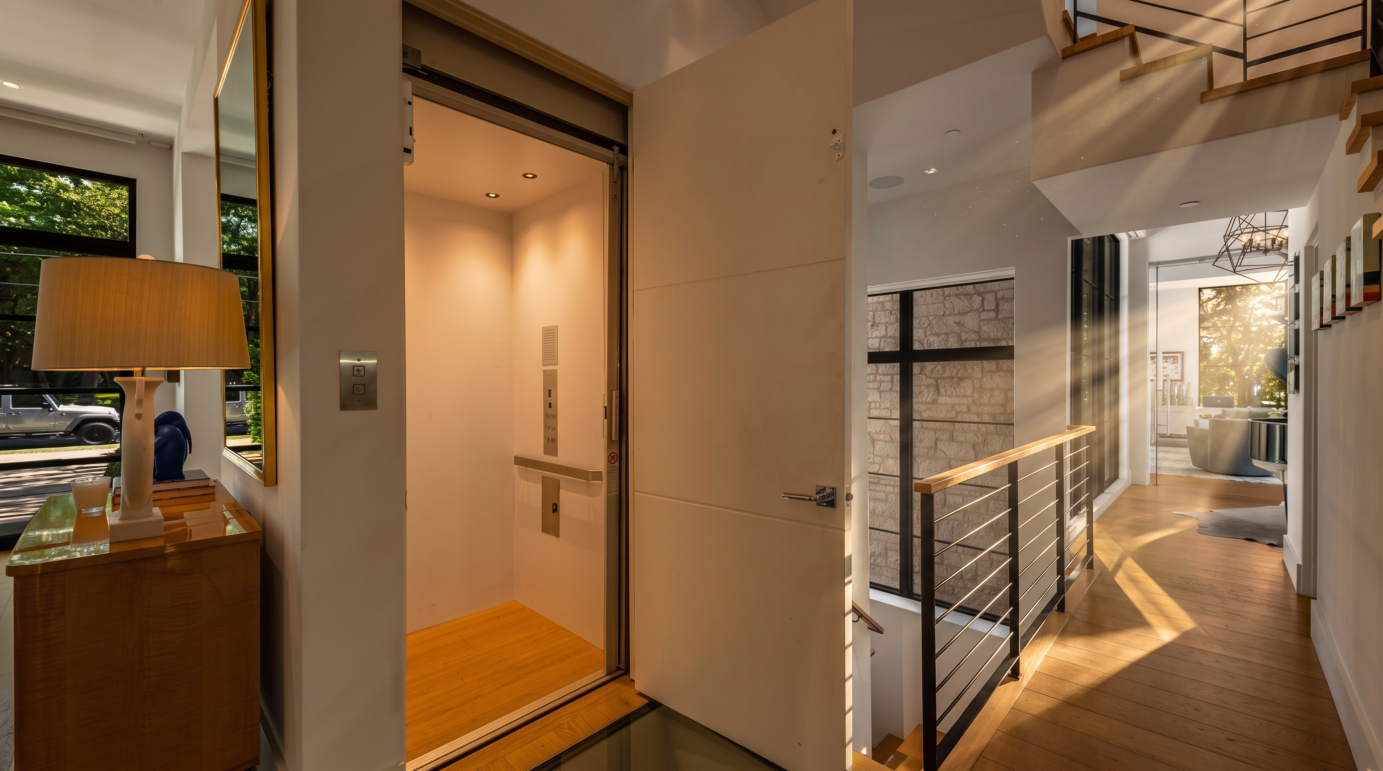 This interior view captures a modern hallway featuring a residential elevator with an open door, positioned next to a staircase with a metal railing. The space is finished with light wood flooring and crisp white walls, creating a bright and airy atmosphere. A console table with a lamp sits to the left, while a glass floor section near the elevator adds a unique architectural detail to the contemporary design.