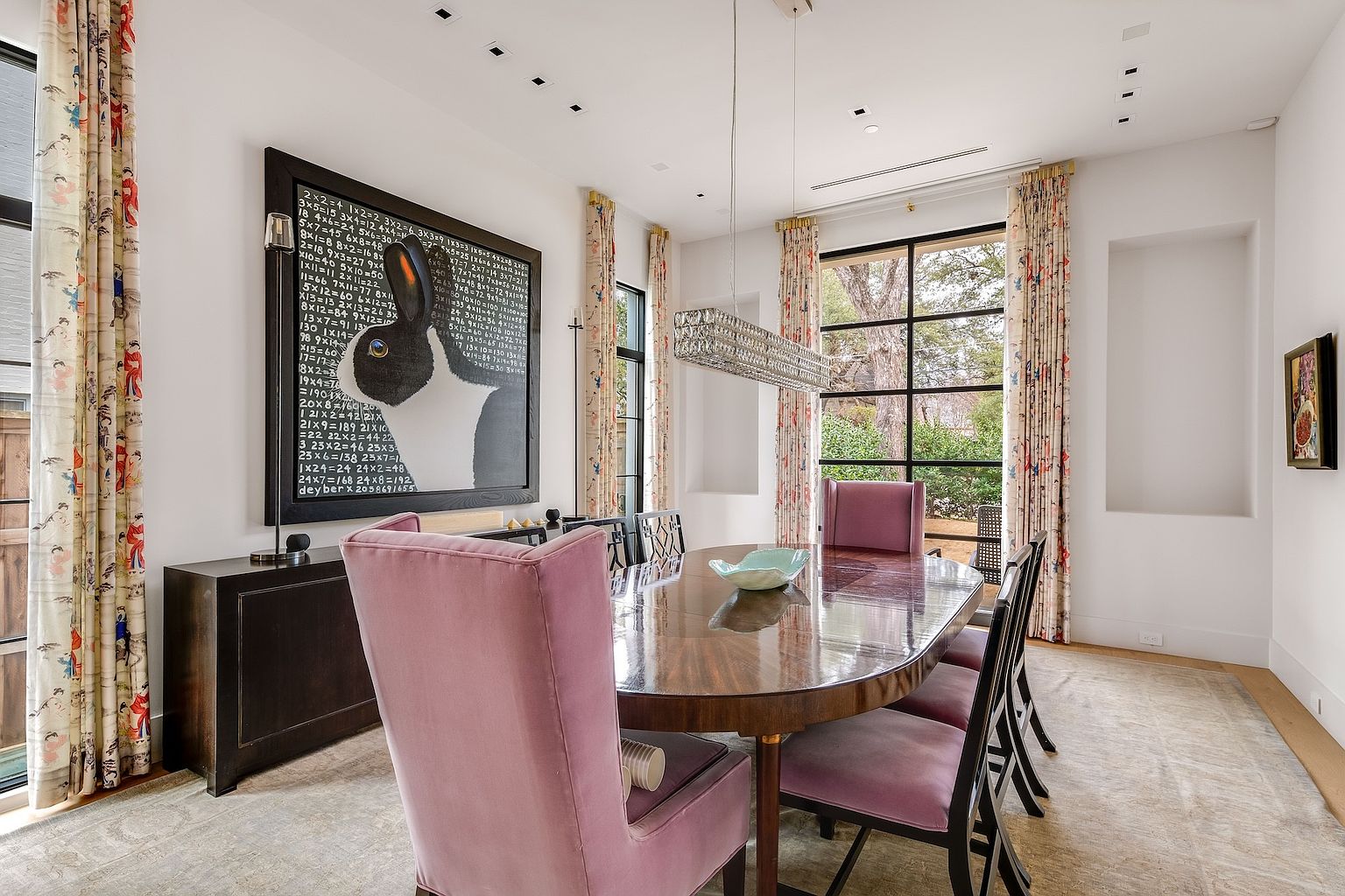 This elegant dining room features a large oval wooden table surrounded by plush, velvet-upholstered chairs in a soft mauve tone. A striking piece of wall art depicting a rabbit against a mathematical backdrop serves as a focal point above a dark sideboard, while a modern, rectangular crystal chandelier hangs above the table. The room is framed by tall windows with patterned curtains, allowing natural light to fill the space and provide a view of the greenery outside.