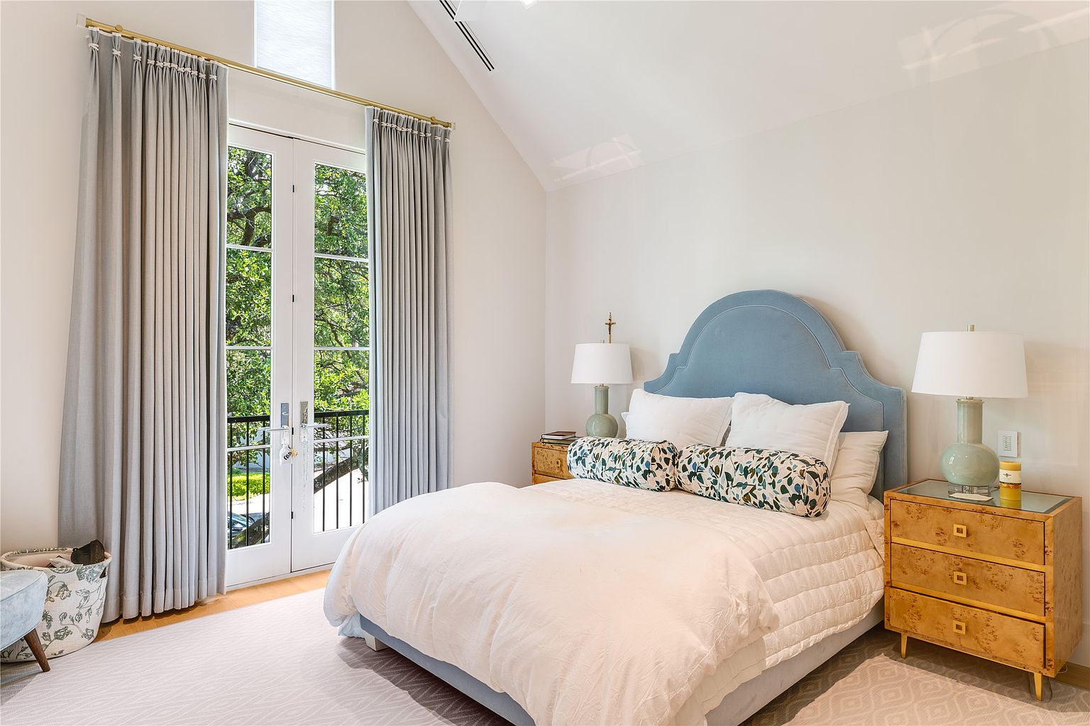 This bright and airy bedroom features a vaulted ceiling and a pair of French doors that open onto a private balcony, allowing for abundant natural light. The room is anchored by a soft blue upholstered headboard, complemented by light-toned wood nightstands and a neutral, textured bedding ensemble. A patterned area rug adds warmth to the space, creating a serene and inviting atmosphere.