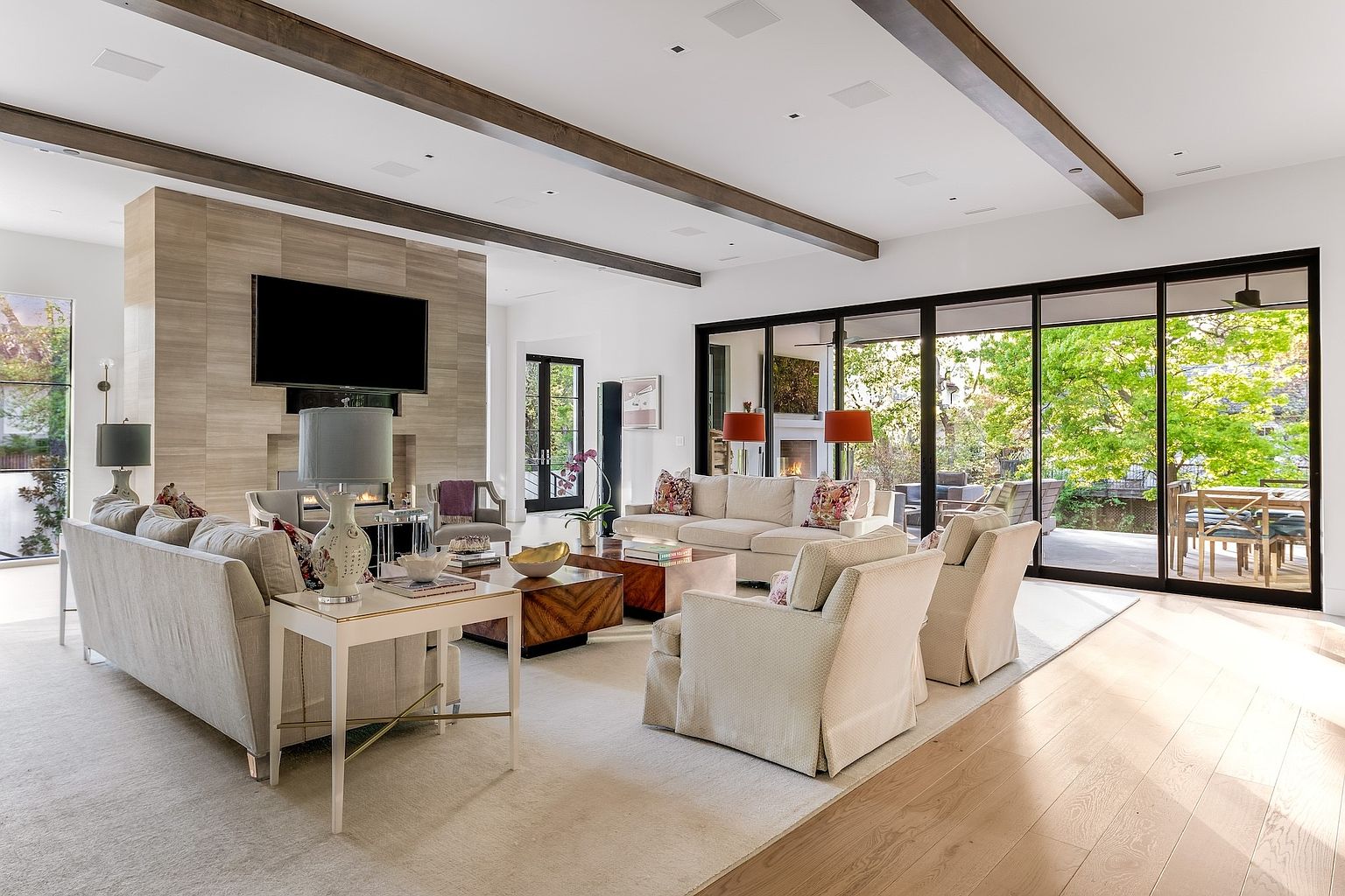 This spacious, light-filled living room features a neutral color palette with plush seating arranged around a central coffee table. A prominent stone-clad chimney breast serves as the focal point, housing a television above a fireplace, while large glass sliding doors provide a seamless transition to the outdoor patio. The room is accented by exposed wooden ceiling beams and warm wood flooring, creating an inviting and sophisticated atmosphere.