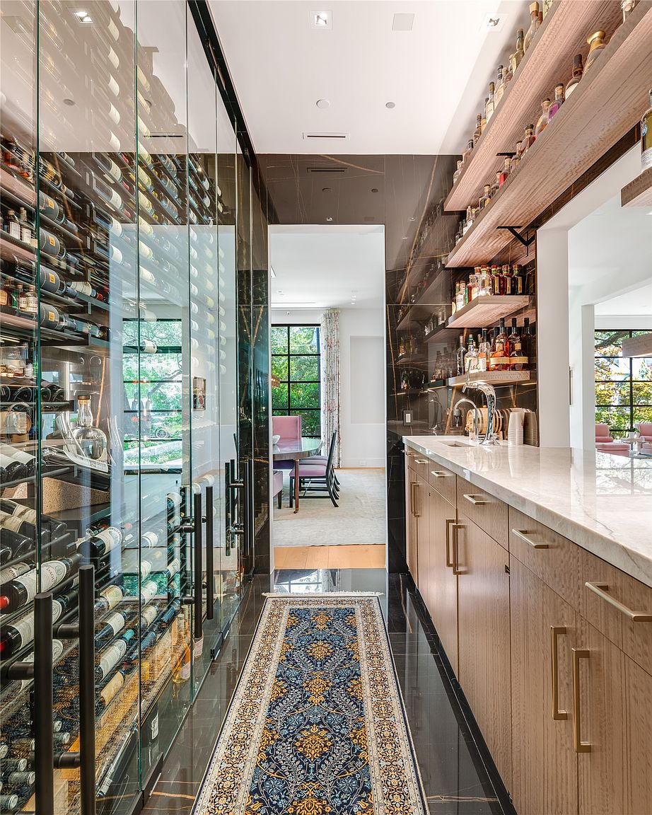 This sophisticated wine cellar and bar area features a glass-enclosed wine storage wall on the left and a sleek bar counter with light wood cabinetry on the right. The space is accented by open shelving for spirits, a dark stone-clad wall, and a long, ornate runner rug that leads the eye toward a bright dining area visible through the doorway. The combination of dark, reflective surfaces and warm wood tones creates a luxurious and modern atmosphere.