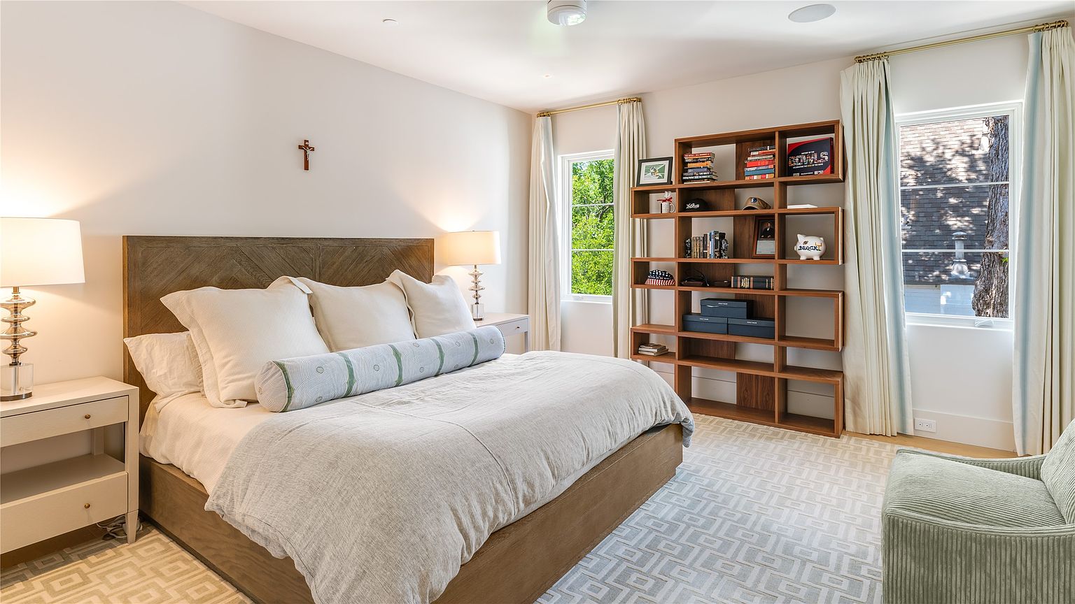 This bright and airy primary bedroom features a large bed with a wood-framed headboard, dressed in neutral linens and a patterned bolster pillow. A tall, open-shelving unit provides ample storage for books and decor, while a patterned area rug adds texture to the space. The room is illuminated by soft natural light from two windows framed by floor-length curtains, creating a serene and inviting atmosphere.
