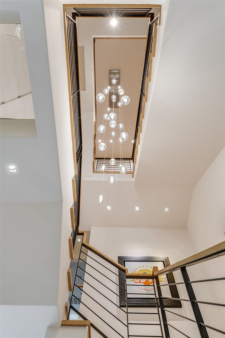 This perspective looks upward through a multi-story stairwell, highlighting a dramatic, cascading glass globe chandelier that serves as the central focal point. The staircase features a modern design with light wood treads, black metal railings, and horizontal cable balusters. A framed piece of artwork hangs on the wall at the lower landing, adding a touch of color to the clean, minimalist aesthetic of the vertical space.