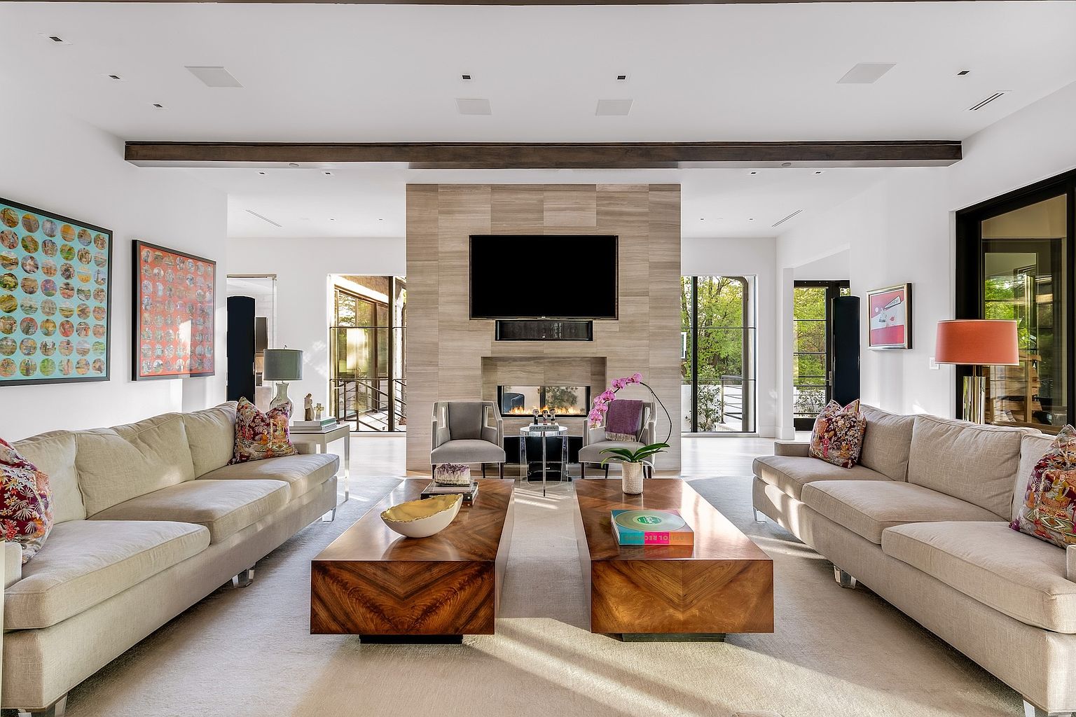 This spacious, symmetrical living room features two matching beige sofas facing each other across a pair of large, dark wood coffee tables. A central stone-clad chimney breast serves as the focal point, housing a television and a fireplace below. The room is framed by dark wood ceiling beams and large windows on either side, creating a bright, balanced, and sophisticated atmosphere.