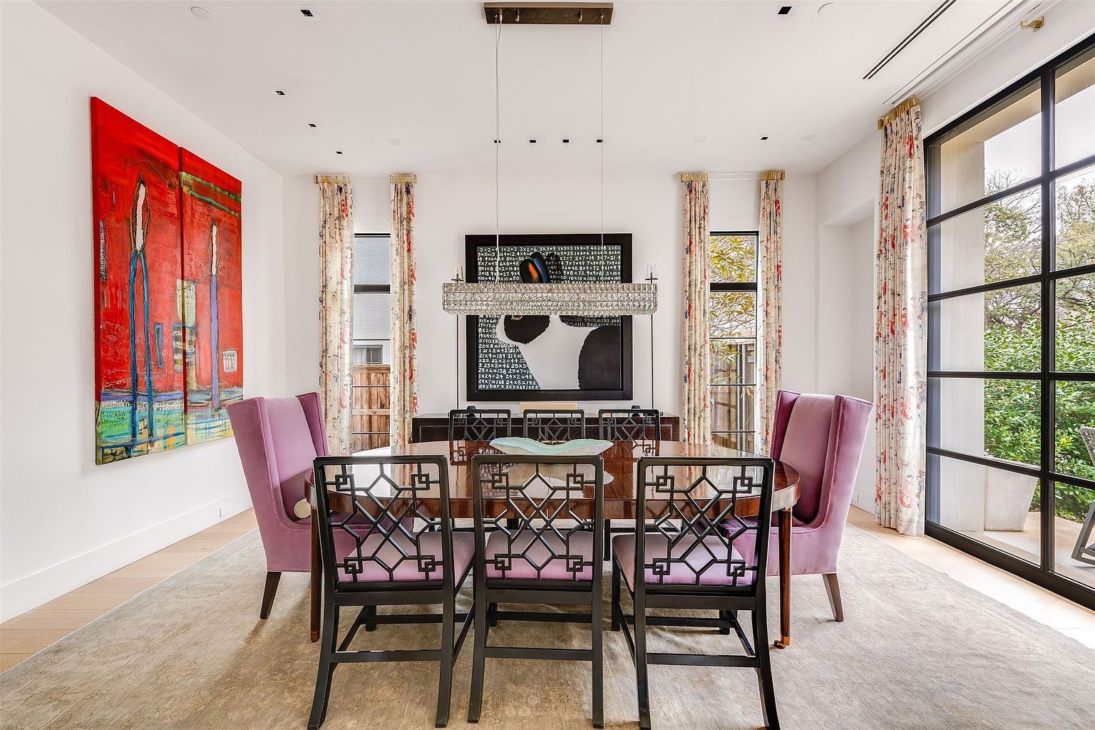 This elegant dining room features a dark wood table surrounded by six chairs, including two upholstered purple armchairs at the heads and four black chairs with geometric backrests. The space is anchored by a large, rectangular crystal chandelier hanging above the table and is framed by tall windows with patterned curtains on either side. A vibrant, large-scale abstract painting hangs on the left wall, while a framed piece of mathematical art serves as a focal point on the back wall above a dark wood sideboard.