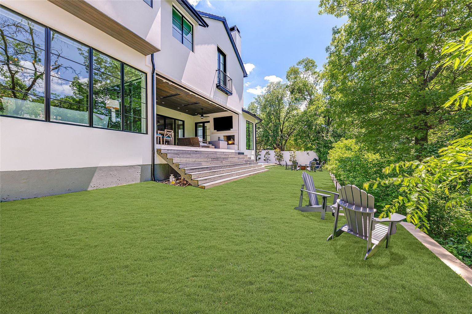 This rear view of a modern home showcases a spacious lawn leading up to a covered patio area with a built-in fireplace and television. The exterior features white siding, large windows, and a set of wide wooden steps that transition from the yard to the outdoor living space. Two Adirondack chairs sit in the foreground, providing a comfortable vantage point to enjoy the lush, tree-lined backyard.
